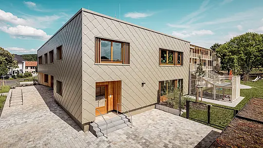 Slika prikazuje gradsku dječju ustanovu u Alzenauu iz dijagonalne ptičje perspektive, ističući modernu arhitekturu s upečatljivom PREFA aluminijskom fasadom u brončanoj boji. Fasada se sastoji od zidnih rombova 44 × 44, koji stvaraju skladan uzorak „ljuski“ i zgradi daju elegantnu dinamiku. Veliki drveni prozori s vertikalnim lamelama unose toplinu i propuštaju obilje dnevnog svjetla u unutrašnjost. S desne strane slike teren se otvara s ograđenim igralištem, zelenim površinama i natkrivenom terasom. Kombinacija dugotrajnog, bezdržavnog aluminija i održivih materijala čini zgradu uzorom funkcionalne i ekološki osviještene arhitekture u obrazovnom sektoru.