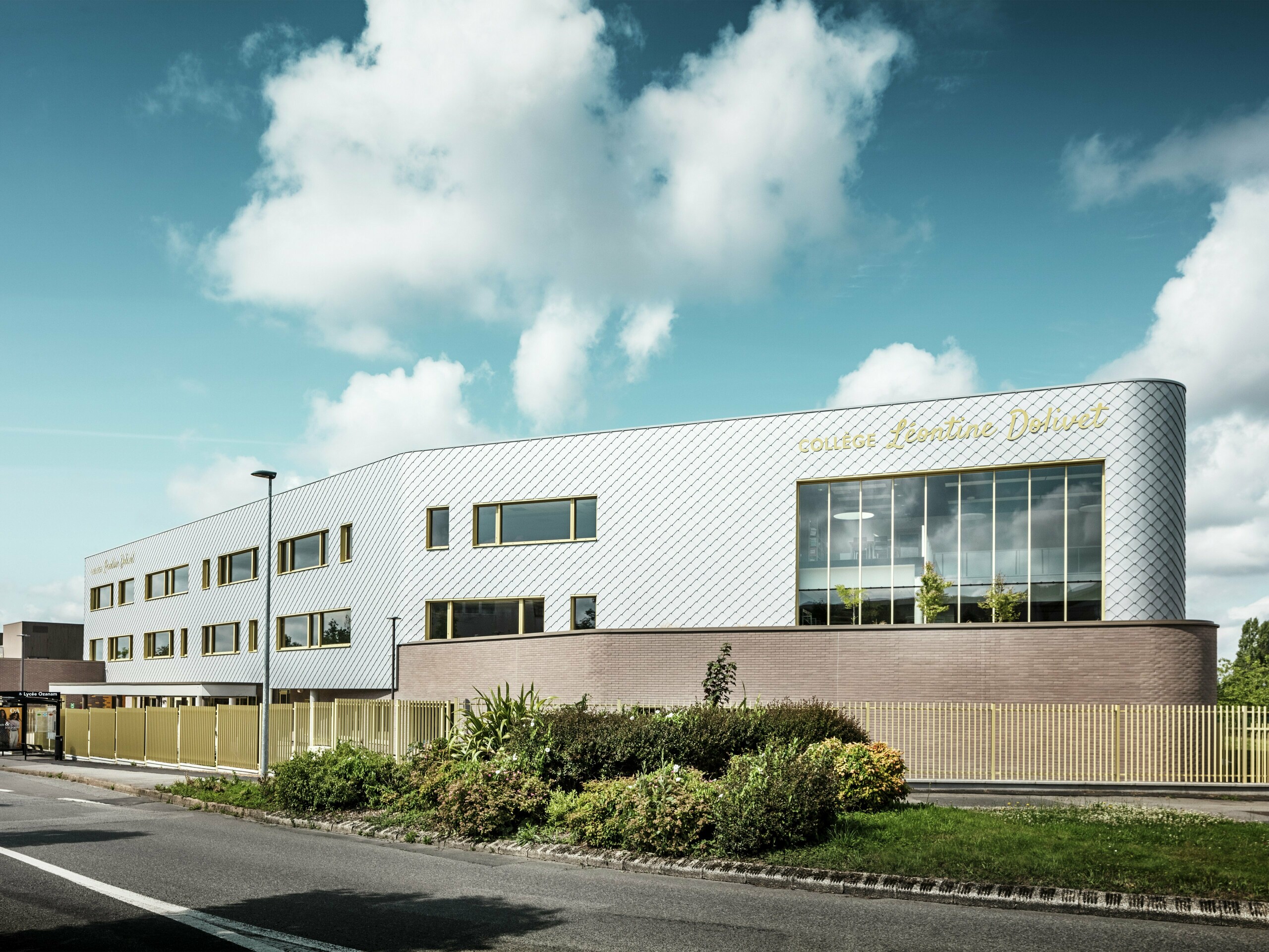 Prikaz Collège Léontine Dolivet s upečatljivom, srebrno sjajnom aluminijskom fasadom od PREFA zidnih rombova 29 × 29 u srebrno-metalik boji. Fasada djeluje dinamično zahvaljujući dijagonalnom uzorku rombova, dok velike staklene površine i zlatni prozorski okviri stvaraju zanimljive kontraste. U prvom planu nalaze se uređene zelene površine i moderni ograda koji uokviruju zgradu. Dugotrajno PREFA rješenje fasade kombinira eleganciju, otpornost na vremenske uvjete i održivost – arhitektonski privlačan objekt u Cesson-Sévignéu, blizu Rennesa.