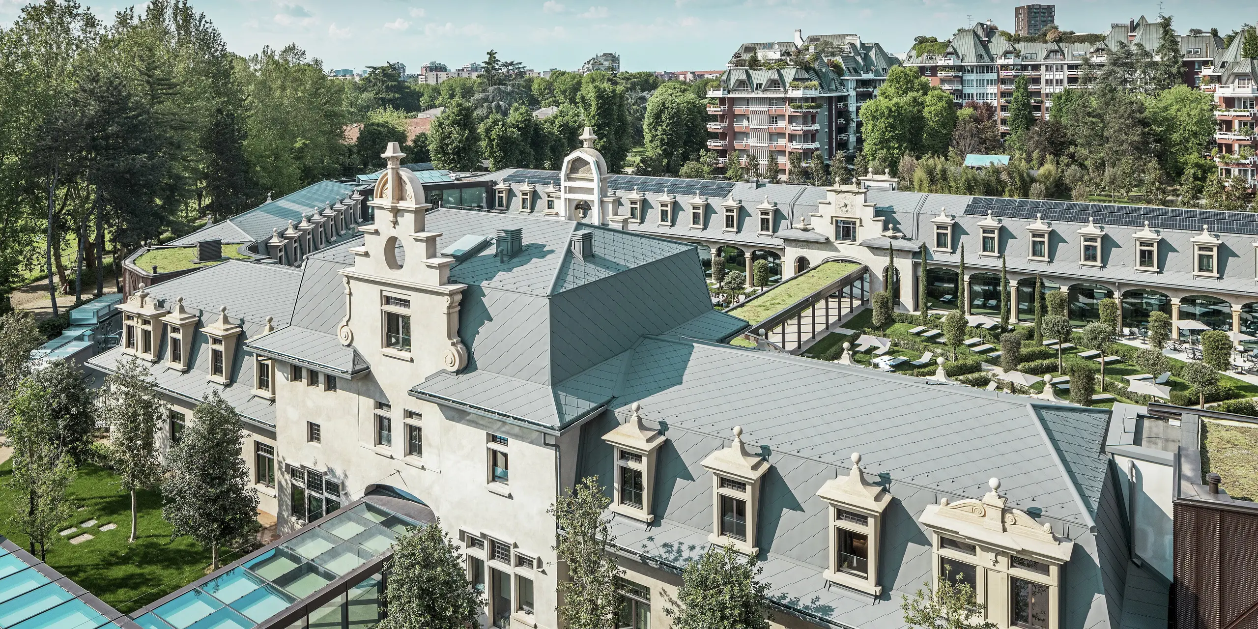 Pogled na impresivan kompleks "De Montel – Terme Milano" s zaštićenom secesijskom arhitekturom i modernim wellness ambijentom. Velika krovna obloga s PREFA krovni romb 44 × 44 u boji P.10 svijetlosiva daje kompleksu elegantnu ujednačenost. Integrirani PV moduli, zeleni krovovi i staklene veze između zgrada odražavaju potražnju za održivost i suvremeni dizajn u urbanom kontekstu.