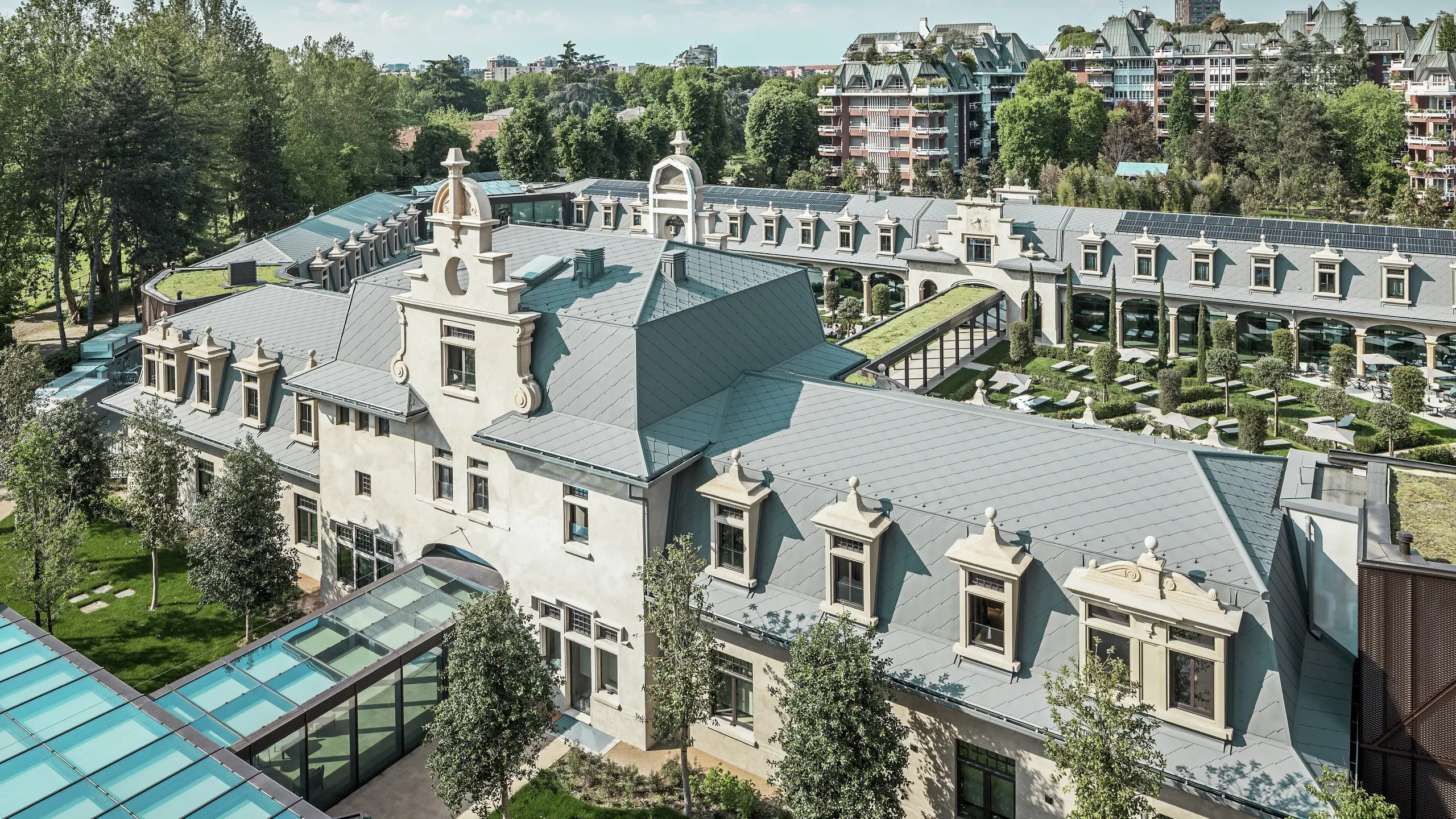Pogled na impresivan kompleks "De Montel – Terme Milano" s zaštićenom secesijskom arhitekturom i modernim wellness ambijentom. Velika krovna obloga s PREFA krovni romb 44 × 44 u boji P.10 svijetlosiva daje kompleksu elegantnu ujednačenost. Integrirani PV moduli, zeleni krovovi i staklene veze između zgrada odražavaju potražnju za održivost i suvremeni dizajn u urbanom kontekstu.