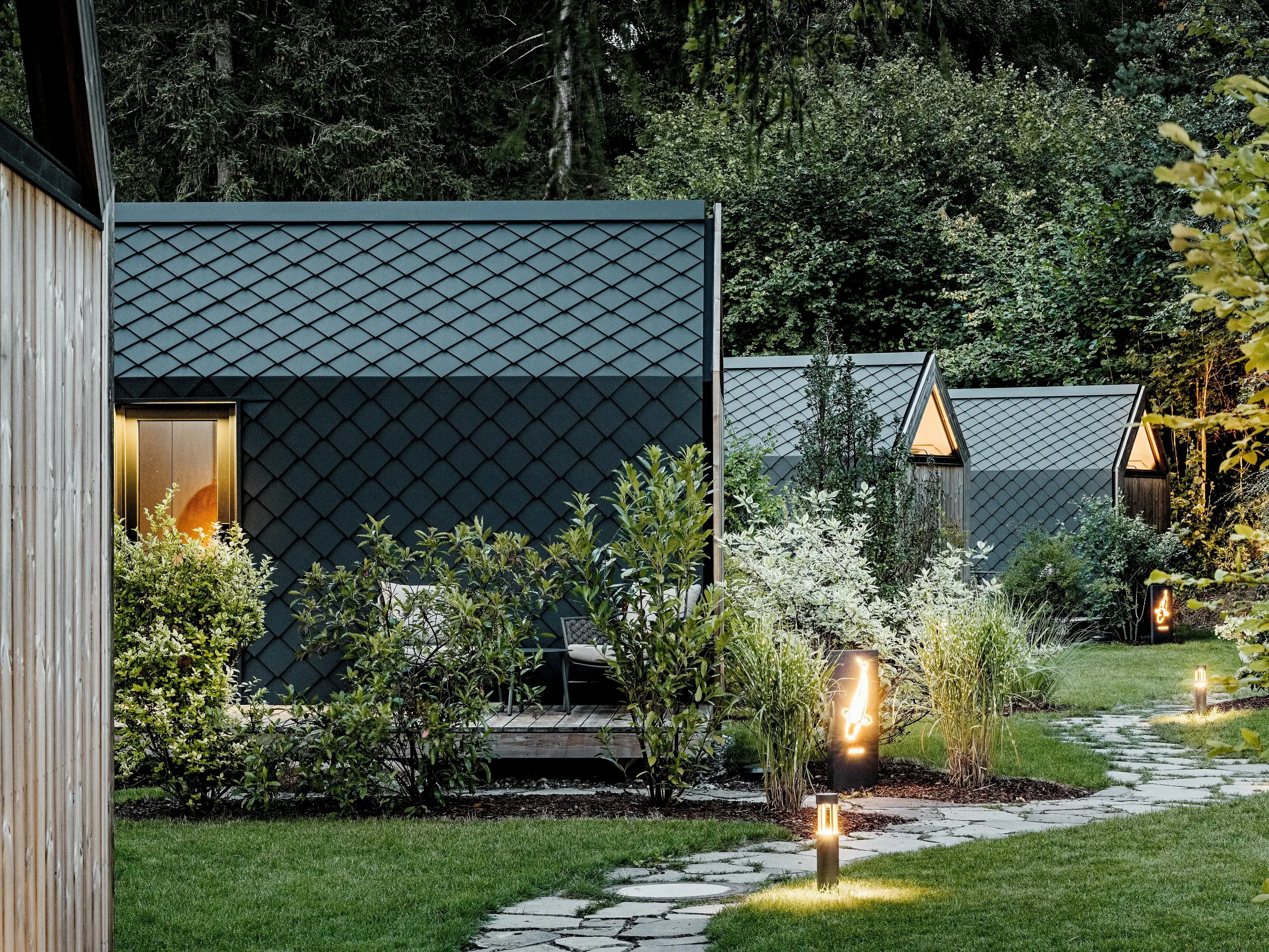 Osvijetljena vrtna staza vodi kroz glamping resort Secret Koi Garden na jezeru Natterer See do nekoliko minimalističkih tiny kuća. Kompaktni volumeni u potpunosti su obloženi PREFA krovnim i zidnim rombovima od aluminija u P.10 antracitnoj boji, čija struktura nalik ribljoj koži u večernjem svjetlu djeluje elegantno.  Uklopljene u bujno zelenilo, trajne i vremenski otporne PREFA aluminijske površine spajaju vrhunsku arhitekturu s prirodom, mirom i održivim dizajnom.
