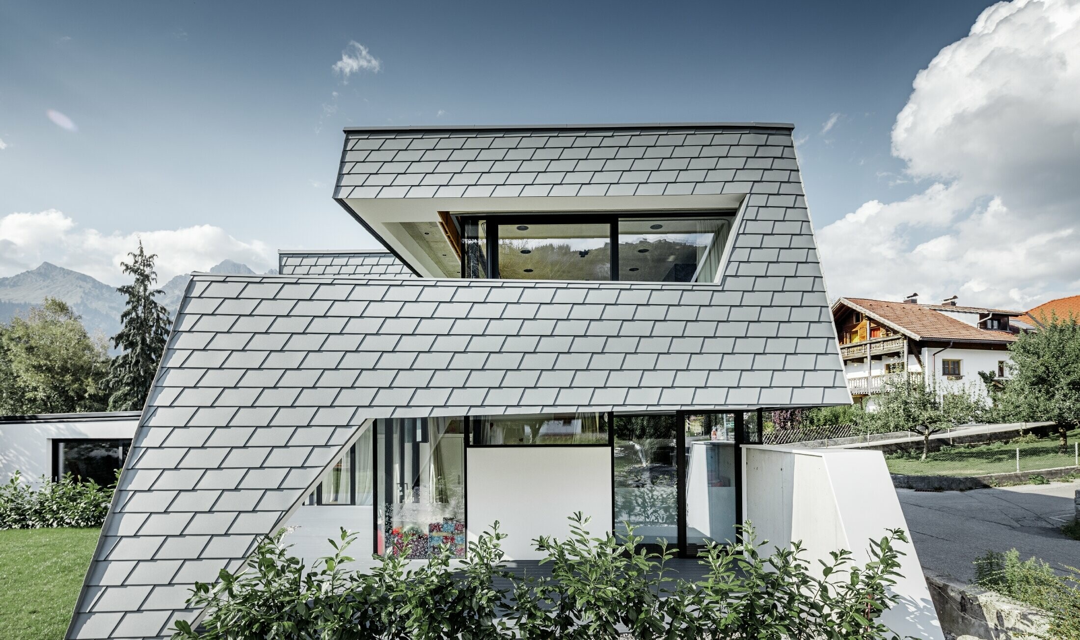 Moderna obiteljska kuća s ravnim krovom, velikim površinama prozora i fasadom iz šindre u svijetlo sivoj boji iz aluminija od PREFE