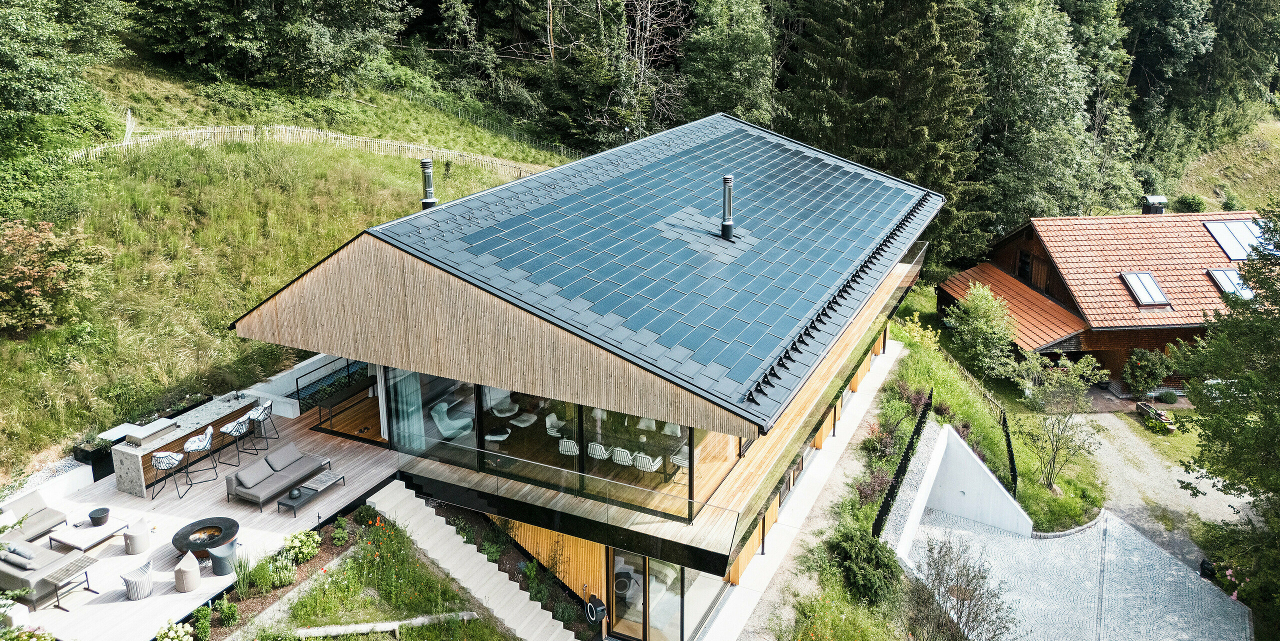 Blick auf die hochwertige Außenküche eines Einfamilienhauses in Egg, Vorarlberg. Das moderne Gebäude wurde mit hochwertigen PREFA Solardachplatten und Dachplatten R.16 in P.10 Schwarz eingedeckt. Das Aluminiumdach mit integrierter Photovoltaikanlage bietet nachhaltige Energiegewinnung und harmoniert mit der einladenden Holzfassade und der umgebenden Natur.