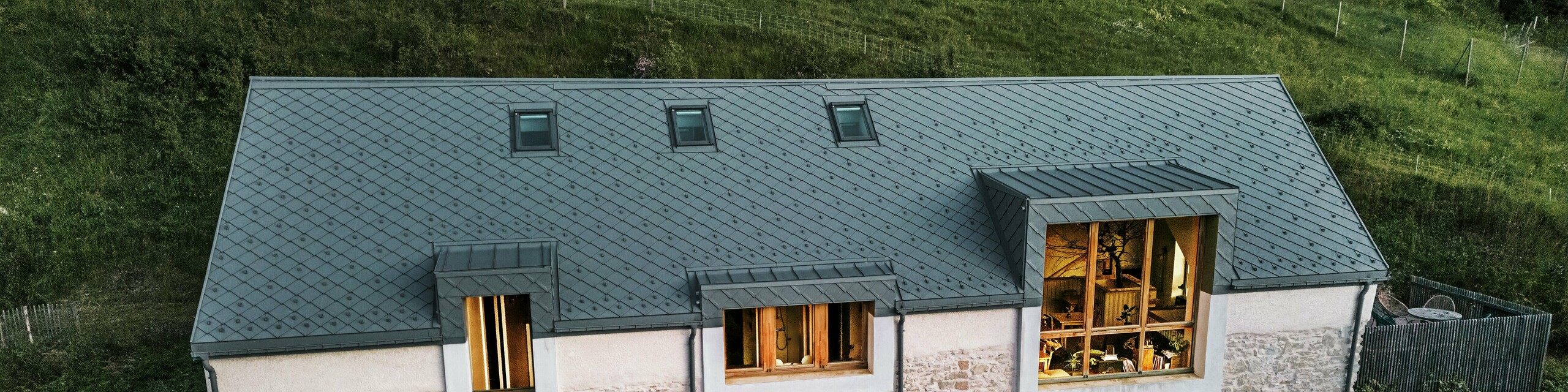 Večernji pogled na obnovljenu štalu "La Grange à Gaby" u Enginsu, Francuska. Zgrada kombinira tradicionalno kameno zidanje s modernim PREFA aluminijskim krovom od rombova veličine 29 × 29 cm. Veliki prozori i osvijetljeni unutarnji prostori naglašavaju ugodnu atmosferu novog kulturnog centra.