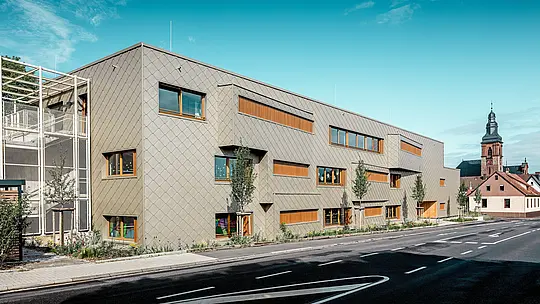U blistavom dnevnom svjetlu moderna dječja ustanova u Alzenauu otkriva se u punoj jasnoći sa svojom izraženom PREFA aluminijskom fasadom od brončano obojenih zidnih rombova 44 × 44. Ravnomjerna „ljuska“ fasade, prekidana pomaknutim prozorima i vertikalnim drvenim lamelama, daje dubinu i ritam cijeloj površini. S lijeve strane slike vidljivo je filigransko stubište s metalnom rešetkastom konstrukcijom, koje zrači transparentnošću i lakoćom. U pozadini se uzdiže kasnogotička crkva u Alzenauu – simbol skladnog povezivanja povijesne arhitekture i suvremenog graditeljstva. Bezdržavno, dugotrajno PREFA fasadno rješenje doprinosi održivoj, funkcionalnoj i estetski visoko kvalitetnoj izvedbi ovog budućnosno orijentiranog obrazovnog objekta.