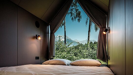 Pogled iz kreveta minimalističke tiny kuće u Secret Koi Gardenu na jezeru Natterer See: kroz veliku trokutastu staklenu frontu prostor se otvara prema prirodi s drvećem i pogledom na tirolski planinski krajolik.  Redukcijski interijer usmjerava fokus prema van i naglašava koncept usporavanja i opuštanja. Arhitekturu štite dugotrajni PREFA aluminijski rombovi koji spajaju dizajn, otpornost na vremenske uvjete i održivost.