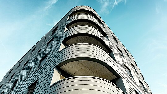 Dinamični prikaz zgrade za stanovanje i poslovanje s upečatljivim zaobljenim kutom i tekućim balkonima. Ljuskasta fasada od PREFA zidnih šindra u P.10 svijetlosivoj boji prekriva zgradu poput precizno obrađene aluminijske kože i naglašava nježne radijuse. Homogena površina ističe jasne linije, dok lagani, otporni na vremenske uvjete PREFA aluminij osigurava najveću oblikovnu stabilnost, dugotrajnost i gotovo bezdržavno održavanje čak i na snažnim zaobljenjima – arhitektonski vizualni naglasak pred sjajno plavim nebom.