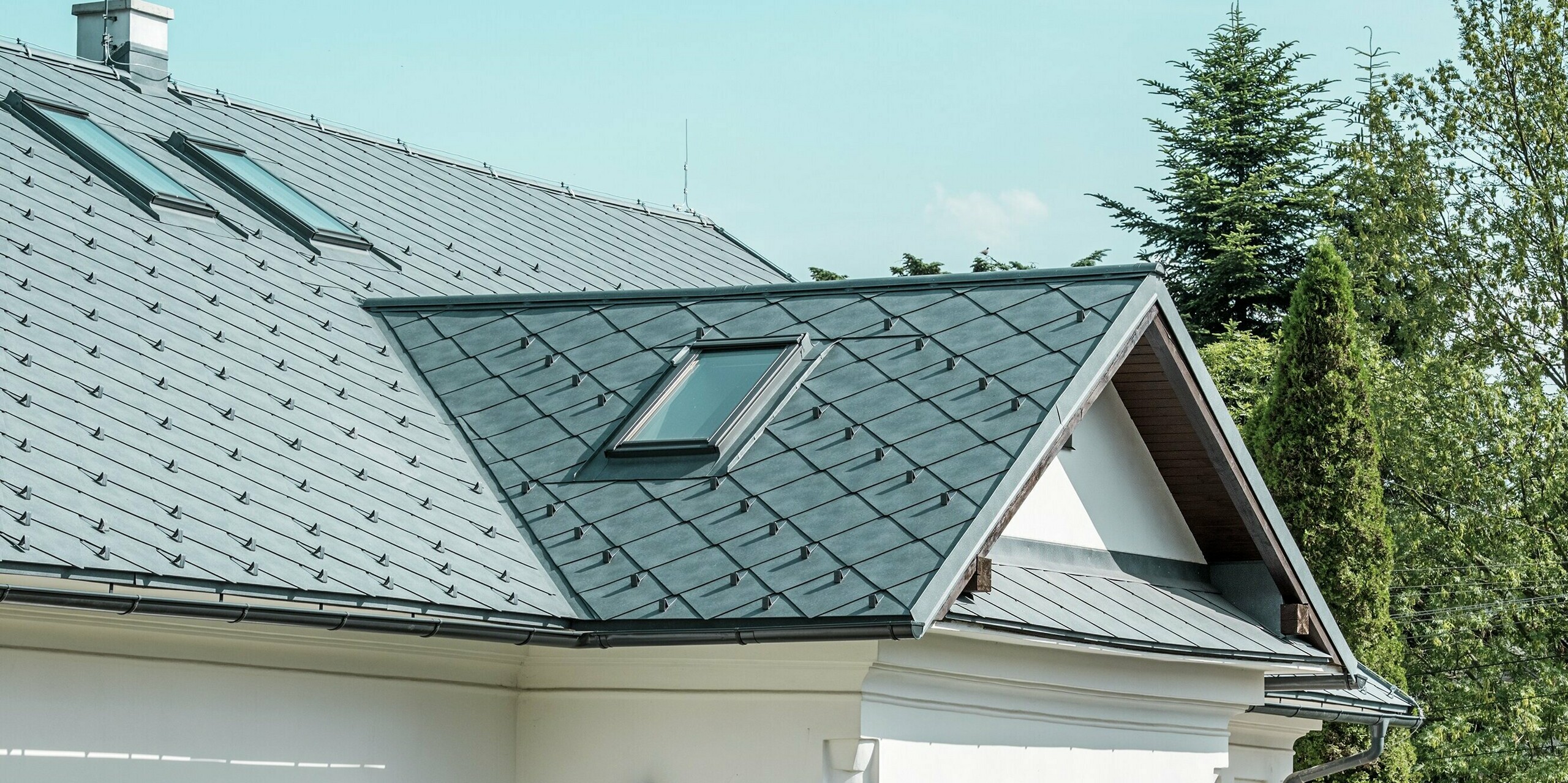 Das Bild zeigt einen detailreichen Ausschnitt des Handwerkerhauses in Klin, Slowakei. Die PREFA Dachraute 44 × 44 in P.10 Steingrau verleiht dem Dach ein modernes, ansprechendes Aussehen und fügt sich perfekt in die traditionelle Architektur des Gebäudes ein. Besonders auffällig sind die großzügigen Dachfenster, die Licht in den oberen Bereich des Hauses bringen und das Design zusätzlich auflockern. Das elegante Satteldach mit der Gaube über dem Eingangsbereich überzeugt mit Langlebigkeit, Leichtigkeit und Widerstandsfähigkeit, was es zur idealen Wahl für eine nachhaltige Sanierung macht.