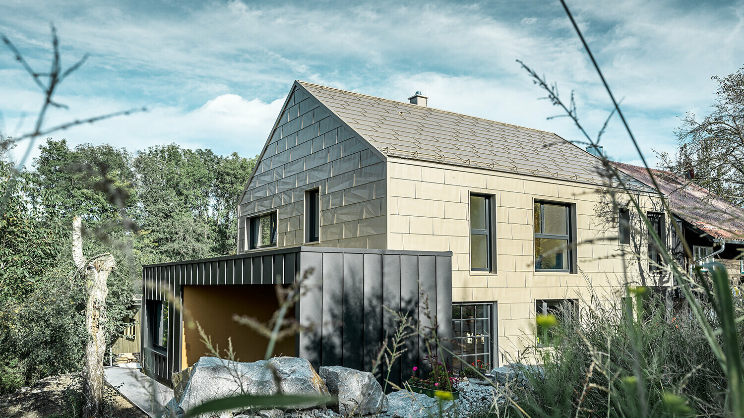 Stambena kuća bez strehe kod koje su krov i fasada obloženi aluminijskim panelom FX.12 tvrtke PREFA u brončanoj boji. Odmah pokraj nalazi se garaža s ravnim krovom s fasadom s kutnim stojećim falcom.