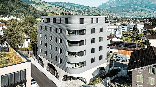 Zračni prikaz Bernerhausa u Buchsu s upečatljivim trokutastim oblikom zgrade i nježno zaobljenim balkonima usred alpskog krajolika. Kontinuirana fasada od PREFA zidnih šindra u P.10 svijetlosivoj boji obavija zgradu poput ljuskaste aluminijske kože i stvara živopisnu igru svjetla i strukture. Iznad masivnog betonskog postolja s lukovima, lagana, otporna na vremenske uvjete PREFA aluminijska fasada djeluje posebno precizno, dugotrajno i gotovo bezdržavno – moderan vizualni naglasak u gradskom okruženju.