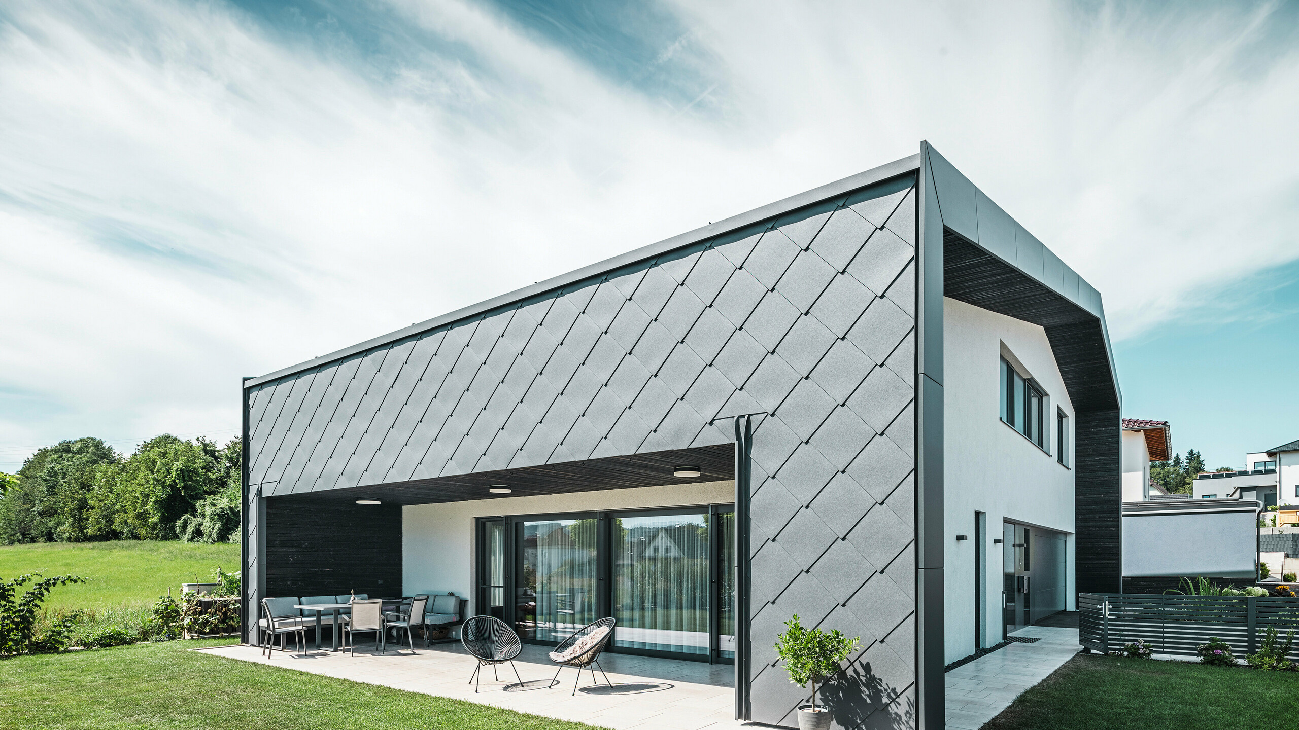 Gepflegter Gartenbereich eines Einfamilienhauses in Aschach. Das Gebäude ist stilvoll eingedeckt mit dunkelgrauen PREFA Aluminium-Wandrauten 44 × 44. Die überdachte Terrasse bietet einen gemütlichen Sitzplatz mit Blick auf die saftig grüne Rasenfläche. Die elegante Aluminiumverkleidung in Kombination mit weißen Putzflächen sorgt für eine moderne und witterungsbeständige Architektur. Große Fensterflächen lassen viel Tageslicht in den Innenraum und schaffen eine offene, lichtdurchflutete Atmosphäre.