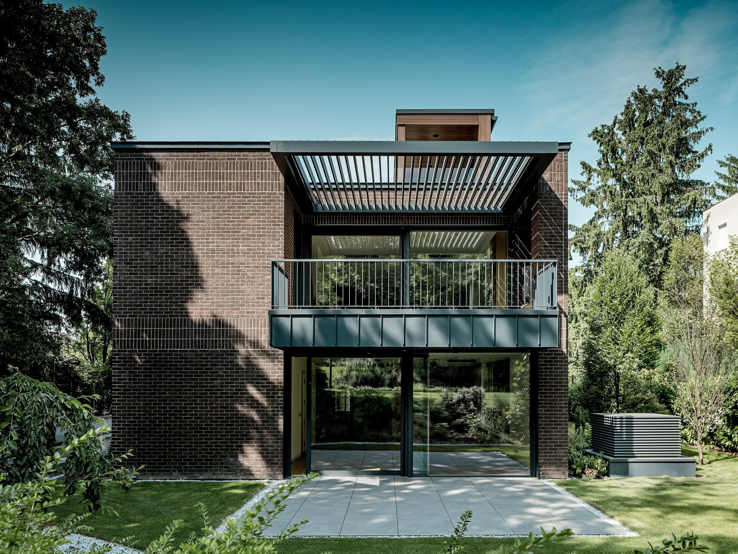 Snimka prikazuje vrtnu stranu moderne višegeneracijske vile u Budimpešti s jasno strukturiranom fasadom od tamnocrvene opeke i PREFALZ aluminija u boji P.10 antracit. Veliki balkon s filigranskom metalnom konstrukcijom i lamelama iznad njega stvara zanimljivu igru svjetla i sjene. Staklene površine od poda do stropa otvaraju interijer prema zelenom vrtu. Precizno izvedeni aluminijski detalji naglašavaju dugotrajnost, otpornost na vremenske uvjete i arhitektonsku jasnoću PREFA rješenja, koja zgradi daju upečatljiv, bezvremenski karakter.