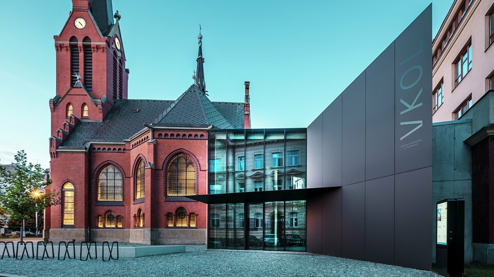 Slika prikazuje impresivnu igru između povijesne Crvene crkve i modernog novog objekta u Olomoucu. Crkva sa svojim karakterističnim gotičkim prozorima i upečatljivim tornjem čini povijesno središte ansambla, dok moderni prizid s elegantnom crnom fasadom od PREFABOND aluminijsko-kompozitnih ploča predstavlja suvremen, arhitektonski odgovor. Velike staklene površine omogućuju pogled u unutrašnjost nove zgrade, gdje se refleksijama u prozorima stvara zanimljiva interakcija između starog i novog. Precizna obrada PREFABOND ploča naglašava modernu estetiku, dok skladna integracija u povijesno okruženje impresionira i funkcionalno i vizualno.