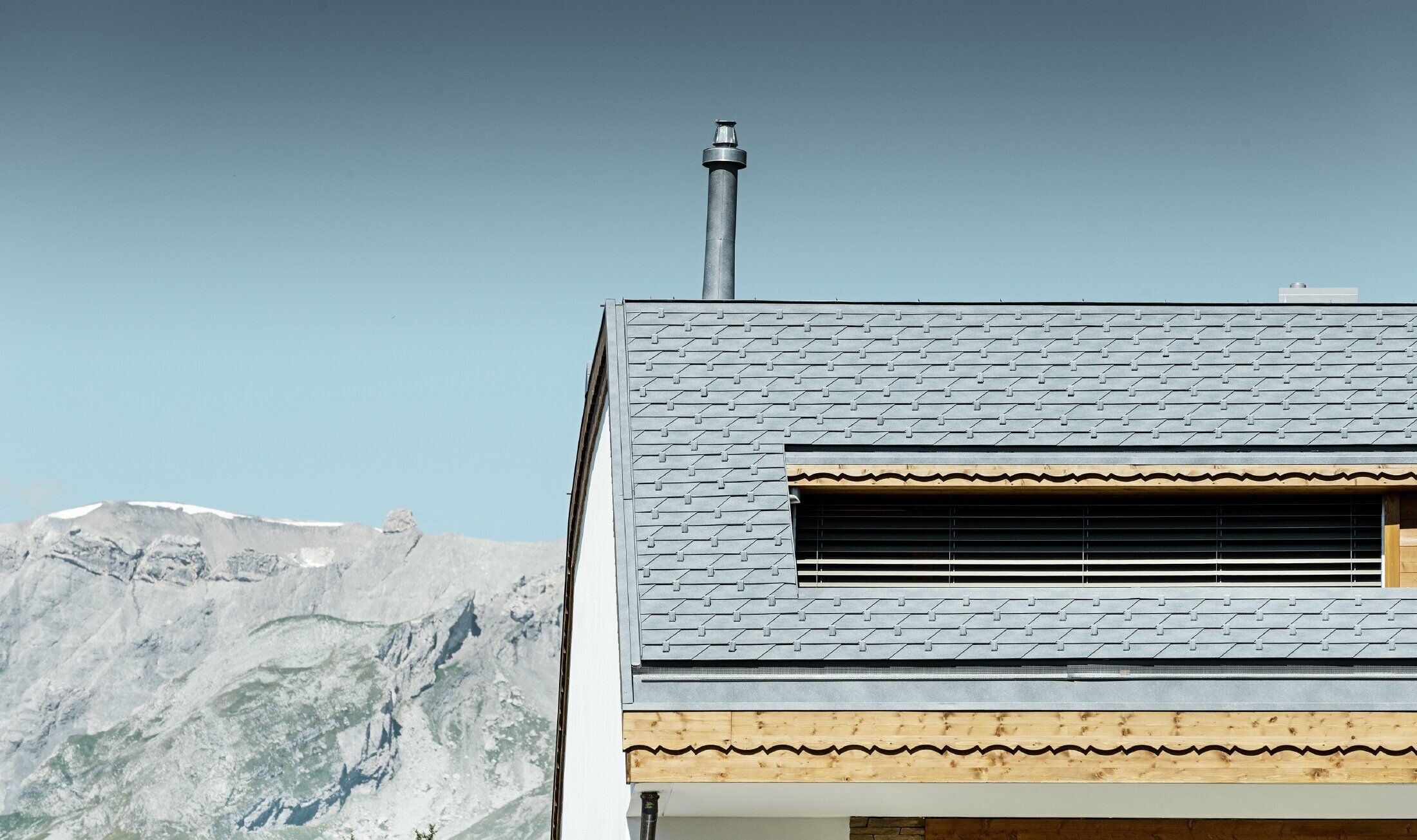 Višeobiteljska kuća u Crans-Montani s brdima u pozadini, s fasadom iz razigranih drvenih elemenata i krovom pokrivenim PREFA aluminijskom krovnom šindrom u kameno sivoj boji