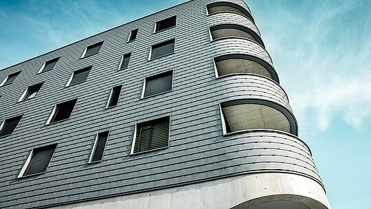 Detaljni prikaz Bernerhausa u Buchsu s upečatljivim zaobljenim kutom zgrade i tekućim otvorima balkona. Fasada od PREFA zidnih šindra u P.10 svijetlosivoj boji prekriva jasno strukturiranu kubaturu poput precizno obrađene aluminijske kože. Ljuskasta površina naglašava nježne radijuse i stvara živopisnu igru svjetla i sjene. Zahvaljujući laganom, otpornom na vremenske uvjete i dugotrajnom PREFA aluminiju, nastaje homogena, gotovo bezdržavna ovojnica zgrade – tehnički zahtjevna, a arhitektonski pravi vizualni naglasak.