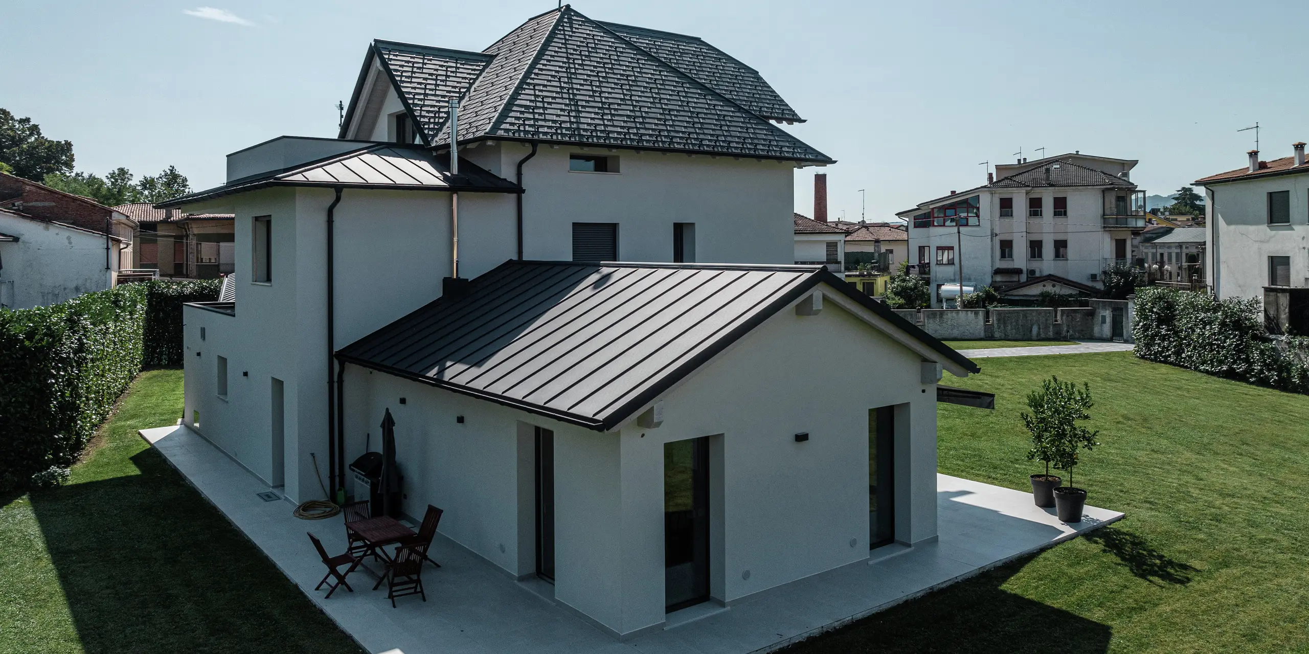 Rückansicht eines zeitgemäßen Einfamilienhauses in Marano Vicentino, Italien, mit einem langlebigen PREFA Aluminiumdach in der hochwertigen P.10-Beschichtung in Anthrazit. Die angrenzenden Nebengebäude sind mit einer robusten Doppelstehfalzdeckung aus Aluminium versehen. Die hochwertige Aluminiumverkleidung verbindet Ästhetik mit Witterungsbeständigkeit.