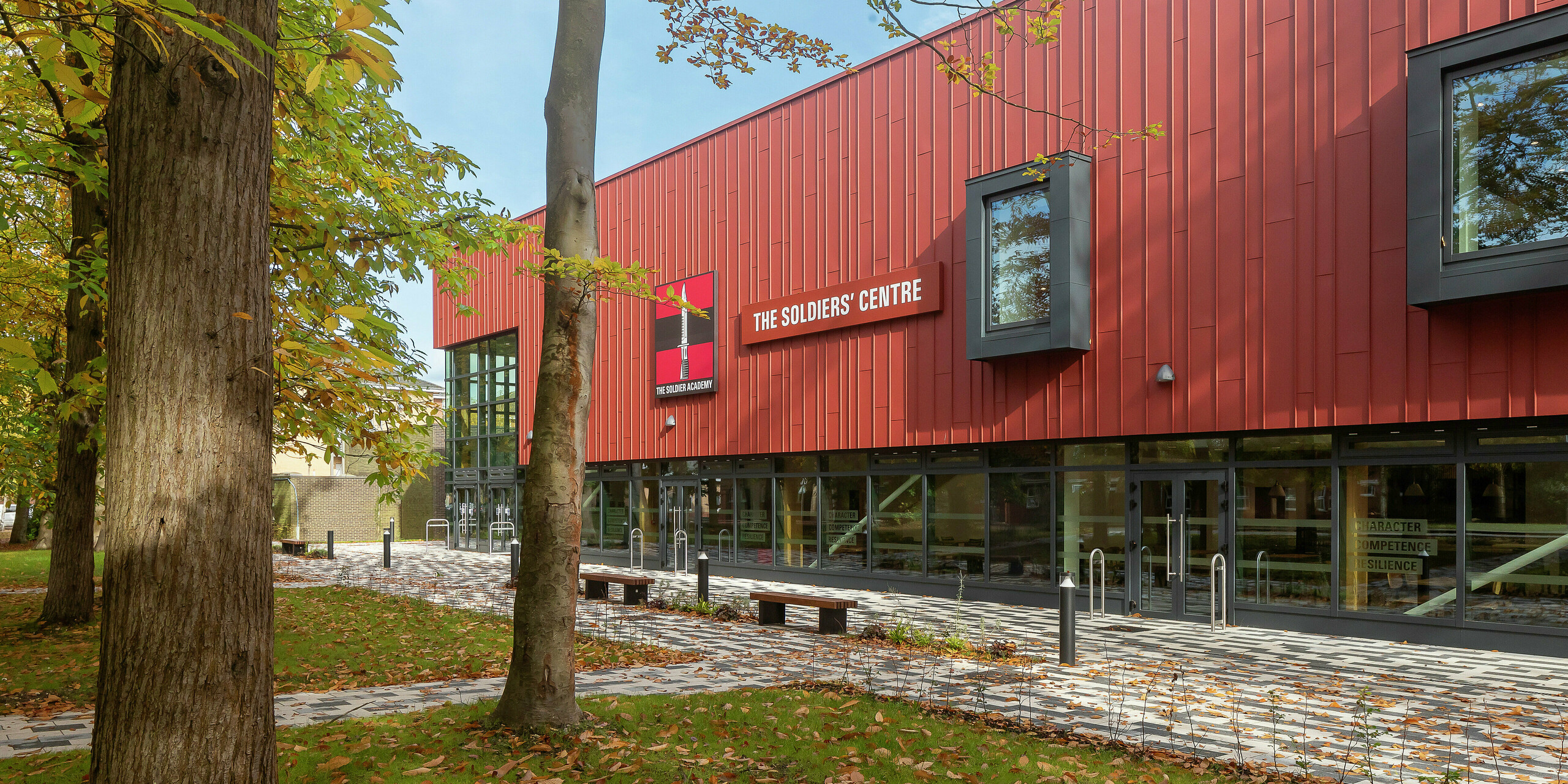 Detailansicht der modernen Fassadenelemente am Soldiers' Centre in Pirbright: Im Mittelpunkt steht die präzise verlegte Stehfalzverkleidung aus PREFALZ in P.10 Oxydrot, die in Kombination mit den markanten Fensterrahmen in P.10 Anthrazit einen optischen Akzent setzt. Die feinen Linien und Schattenspiele unterstreichen die hochwertige Verarbeitung und das innovative Design der PREFA Aluminiumprodukte - ein Beweis für nachhaltige und zeitgemäße Architektur.