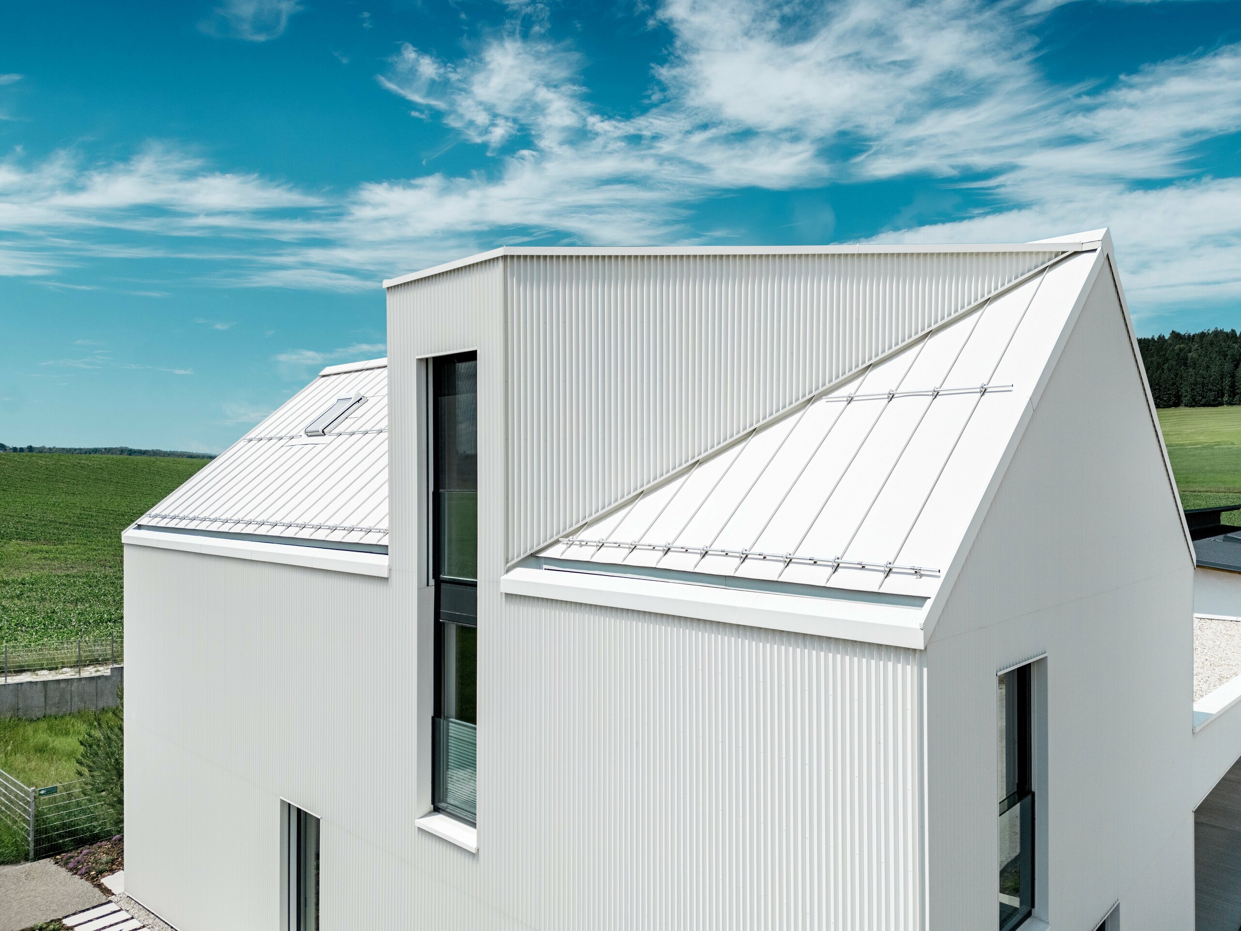 Slika prikazuje strmi, upečatljivi zabatni krov ove obiteljske kuće u Freistadtu, prekriven PREFALZ-om u boji čistobijele. Precizno naborana fasada i krov besprijekorno su povezani, stvarajući elegantne, čiste linije. PREFALZ korišten za krov i fasadu osigurava ne samo estetsku dugovječnost, već i visoku otpornost na vremenske uvjete. Time zgrada dobiva dugoročnu izdržljivost i jednostavno održavanje. Kombinacija modernog dizajna i funkcionalnih materijala stvara skladnu, suvremenu arhitekturu koja je i praktična i vizualno privlačna. Posebno upada u oči krovni prozor s kosim nagibom i prozorom koji se proteže kroz dvije etaže.