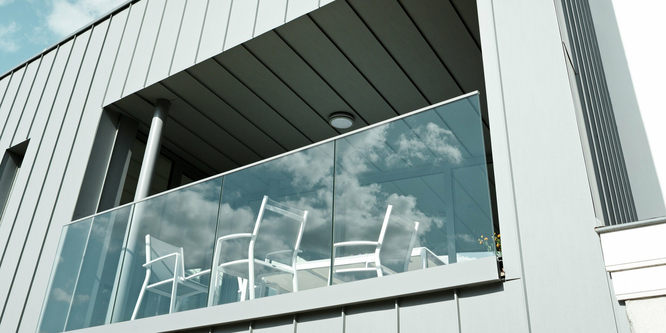 Stilvolle Detailaufnahme einer überdachten Terrasse mit Glasbrüstung an einer Fassade aus PREFALZ in Patinagrau. Die vertikale Stehfalzstruktur aus Aluminium verleiht dem Gebäude klare Linien und ist zudem witterungsbeständig, pflegeleicht und langlebig – ideal für anspruchsvolle Architektur.
