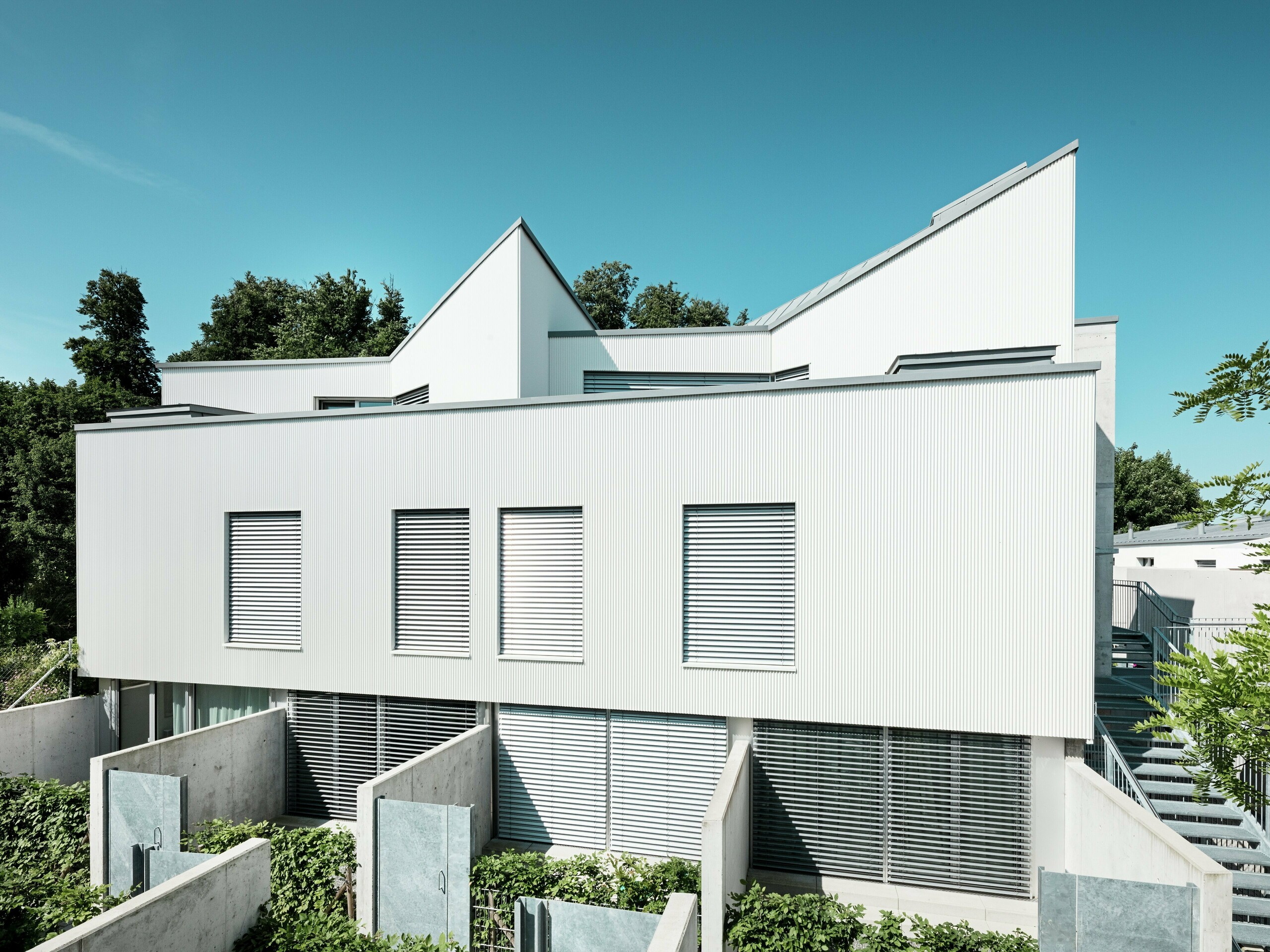 Suvremeni Tiny Homes na Zollikerbergu u Švicarskoj impresioniraju čistim linijama i modernom PREFA aluminijskom fasadom od ekstruzijskih zubčastih profila. Vertikalno postavljeni, nježno zakrivljeni profili stvaraju upečatljivu i elegantnu fasadu koja oživljava zahvaljujući refleksiji svjetla. Bijela površina daje dojam lakoće i naglašava geometrijsku arhitekturu. Predmontirana, ventilirana fasada trajno štiti zgradu od vremenskih utjecaja i poboljšava klimu prostora. Površina je lako održiva i otporna na koroziju.