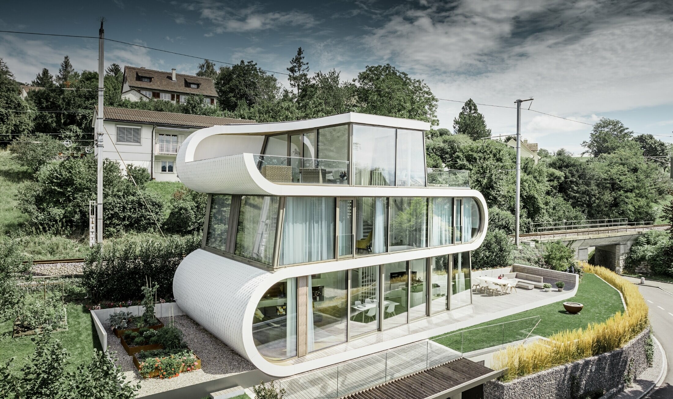 Vrlo moderna obiteljska kuća koju je dizajnirao arhitekt Camenzind iz Züricha; Vijugava traka međusobno spaja pojedine razine. Zaobljenja su obložena malim PREFA krovnim rombom u bijeloj boji. Kuća ima mnoge i velike staklene površine zbog čega djeluje vrlo otvoreno.