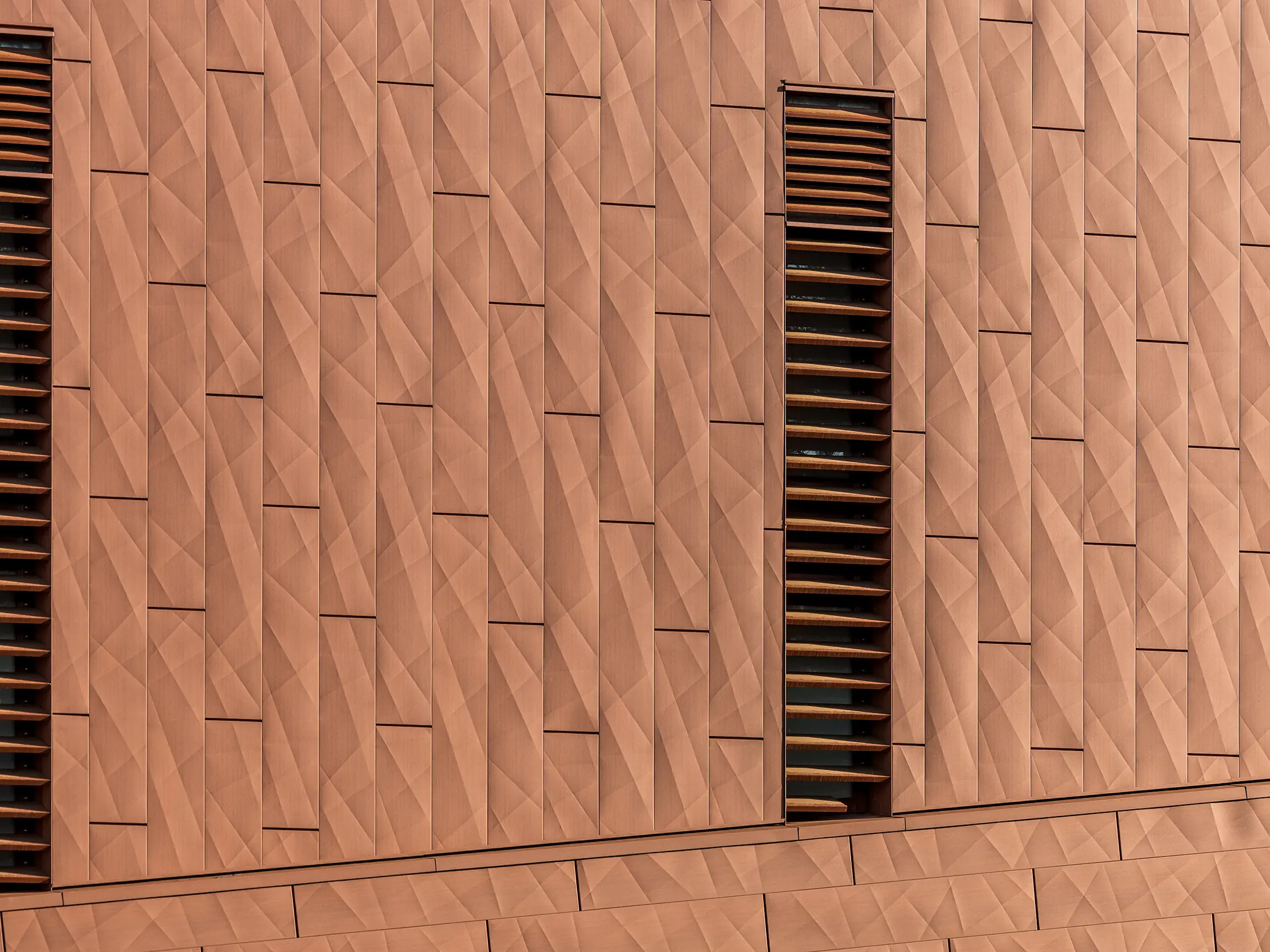 Detaljni prikaz PREFA Siding.X aluminijske fasade na postrojenju za pitku vodu WML u Lottumu s fino presavijenom, geometrijskom površinom u toplom izgledu hrđe. Precizno raspoređeni paneli stvaraju živahnu igru svjetla i sjene, dok vitki, vertikalni lamelni otvori kao upečatljivi vizualni elementi strukturiraju izgled fasade. Lagani, dugotrajni i potpuno reciklabilni PREFA aluminijski paneli odlikuju se visokom otpornošću na vremenske uvjete, postojanošću boje te održivim fasadnim omotačem koji zahtijeva minimalno održavanje.