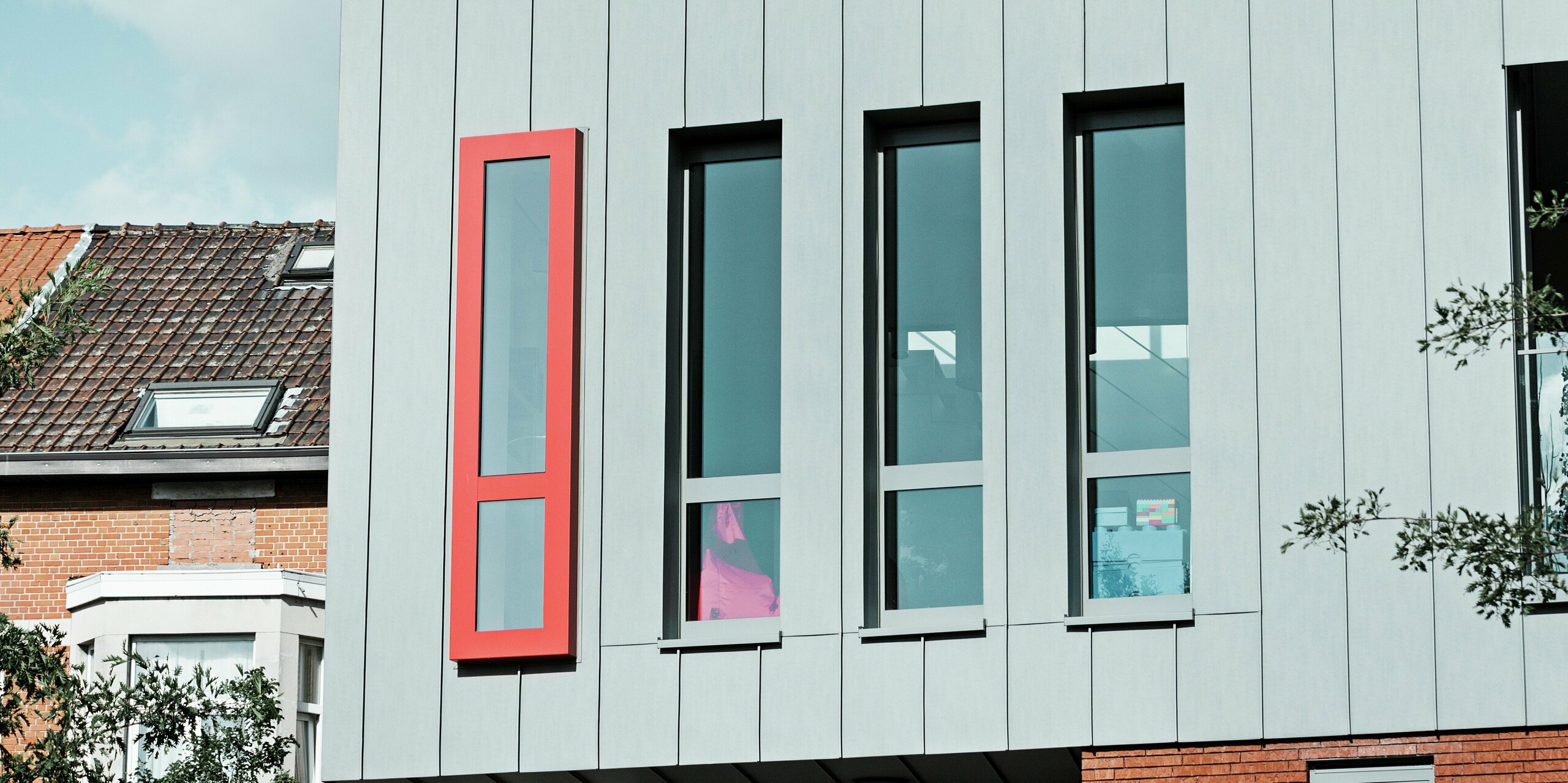 Diese Detailaufnahme zeigt die moderne PREFALZ-Aluminiumfassade in der Farbe Patinagrau mit vertikaler Stehfalzstruktur. Besonders ins Auge fällt das schmale, hochformatige Fenster mit dem kräftig roten Fensterrahmen, der einen architektonischen Akzent setzt. Die großzügigen Glasflächen sorgen für Licht im Gebäude. 