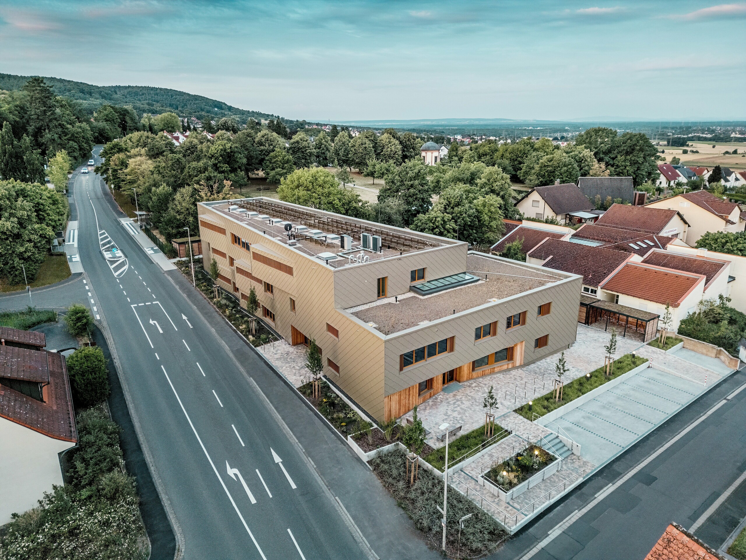 Impresivni ukupni dojam gradske dječje ustanove u Alzenauu postaje vidljiv iz zračne snimke. L‑oblik hibridne zgrade skladno je smješten u mirnu stambenu četvrt na rubu Spessarta. Fasada od brončano obojenih PREFA zidnih rombova 44 × 44 daje građevini upečatljiv, visokokvalitetan izgled i naglašava modernu arhitekturu. Zeleni krovovi, prostrani vanjski prostori i jasna organizacija prometa dodatno ističu održivi koncept cijelog projekta. Kombinacija dugotrajnog aluminija, drva i recikliranog betona zadovoljava najviše ekološke standarde. Ova perspektiva ističe promišljenu urbanističku integraciju i multifunkcionalni karakter zgrade – budućnosno orijentiran projekt za obrazovanje, stanovanje i održivost.