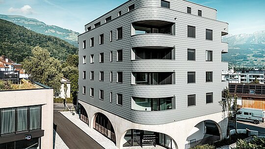 Vanjski prikaz Bernerhausa u Buchsu s upečatljivim, trokutastim oblikom i nježno zaobljenim balkonima. Fasada od PREFA zidnih šindra u P.10 svijetlosivoj boji obavija zgradu poput ljuskaste aluminijske kože i stvara preciznu, živopisnu fasadnu kompoziciju. Iznad masivnog betonskog postolja s elegantnim lukovima, lagana, otporna na vremenske uvjete PREFA aluminijska fasada djeluje posebno izražajno. Dugotrajna, gotovo bezdržavna i oblikovno stabilna, postavlja moderno arhitektonsko istaknuto mjesto pred alpskim planinskim krajolikom.