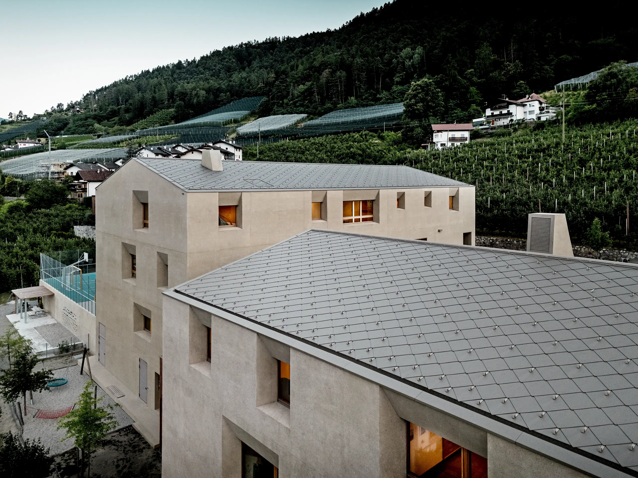 Iz ptičje perspektive obrazovni centar Riffian u Južnom Tirolu pokazuje svoje prepoznatljive PREFA krovne rombove 44 × 44 u boji P.10 bronca. Precizno izvedena aluminijska krovna obloga skladno se proteže preko dječjeg vrtića i osnovne škole, pružajući pouzdanu i dugotrajnu zaštitu od vremenskih utjecaja te dojmljivo uklapajući modernu arhitekturu u voćnjake i obronke planina.
