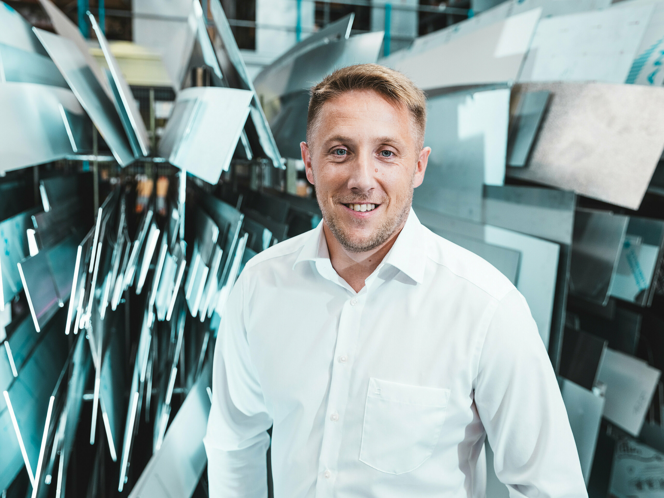 Samouvjeren i prijateljski stoji voditelj projekta Marcel Schmidt iz limarske radionice ASA Schüßler ispred impresivnog zida sastavljenog od aluminijskih elemenata različitih oblika. Dinamično raspoređene metalne ploče efektno reflektiraju svjetlo i simboliziraju inovativnost, preciznost i kvalitetu u obradi metala. Čovjek u bijeloj košulji utjelovljuje stručnost i suvremenu izvrsnost proizvodnje. U pozadini se jasno vidi raznolikost primjene aluminija – od krovnih i fasadnih sustava do specijalno izrađenih rješenja po mjeri.