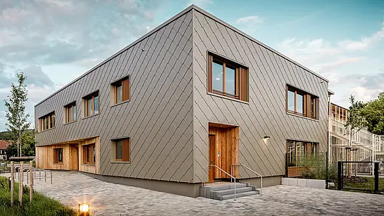 Moderni dječji vrtić u Alzenauu impresionira svojom jasnom arhitekturom i visokokvalitetnom PREFA aluminijskom fasadom u brončanoj boji. Fasada od zidnih rombova 44 × 44 daje zgradi elegantan, strukturiran izgled i vizualno povezuje različite funkcionalne prostore u skladnu cjelinu. Drvene obloge na ulazima stvaraju topao, prirodan kontrast dugotrajnom aluminiju. Kombinacija masivne konstrukcije, drvenih elemenata i ventilirane, prednje postavljene fasade primjer je održive gradnje. Održiva PREFA „odora“ zgrade impresionira ne samo reciklabilnošću, već i otpornošću na vremenske uvjete – idealno za budućnosno orijentirane obrazovne objekte s visokim arhitektonskim standardima.