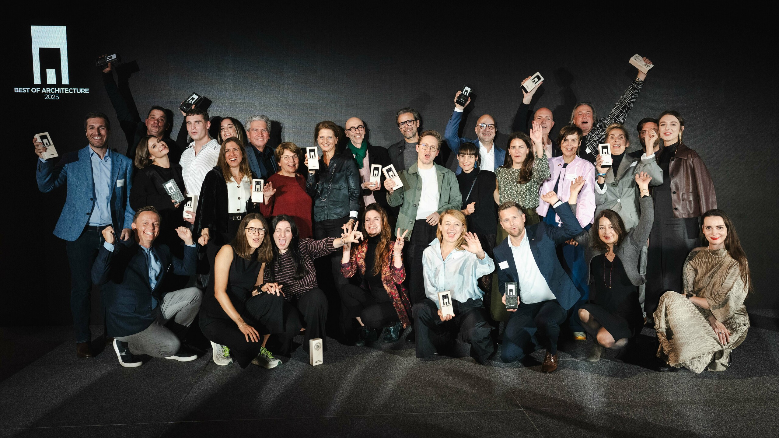 Skupna fotografija pobjednika na svečanosti dodjele nagrade "Stambena zgrada godine 2025": Pobjednici stoje ispred elegantne crne pozornice s logotipom "Best of Architecture" i ponosno drže svoje nagrade u rukama. Dinamične poze, moderna odjeća i upečatljivo osvjetljenje naglašavaju važnost događaja, dok pozornica predstavlja inovaciju i arhitektonsku izvrsnost.