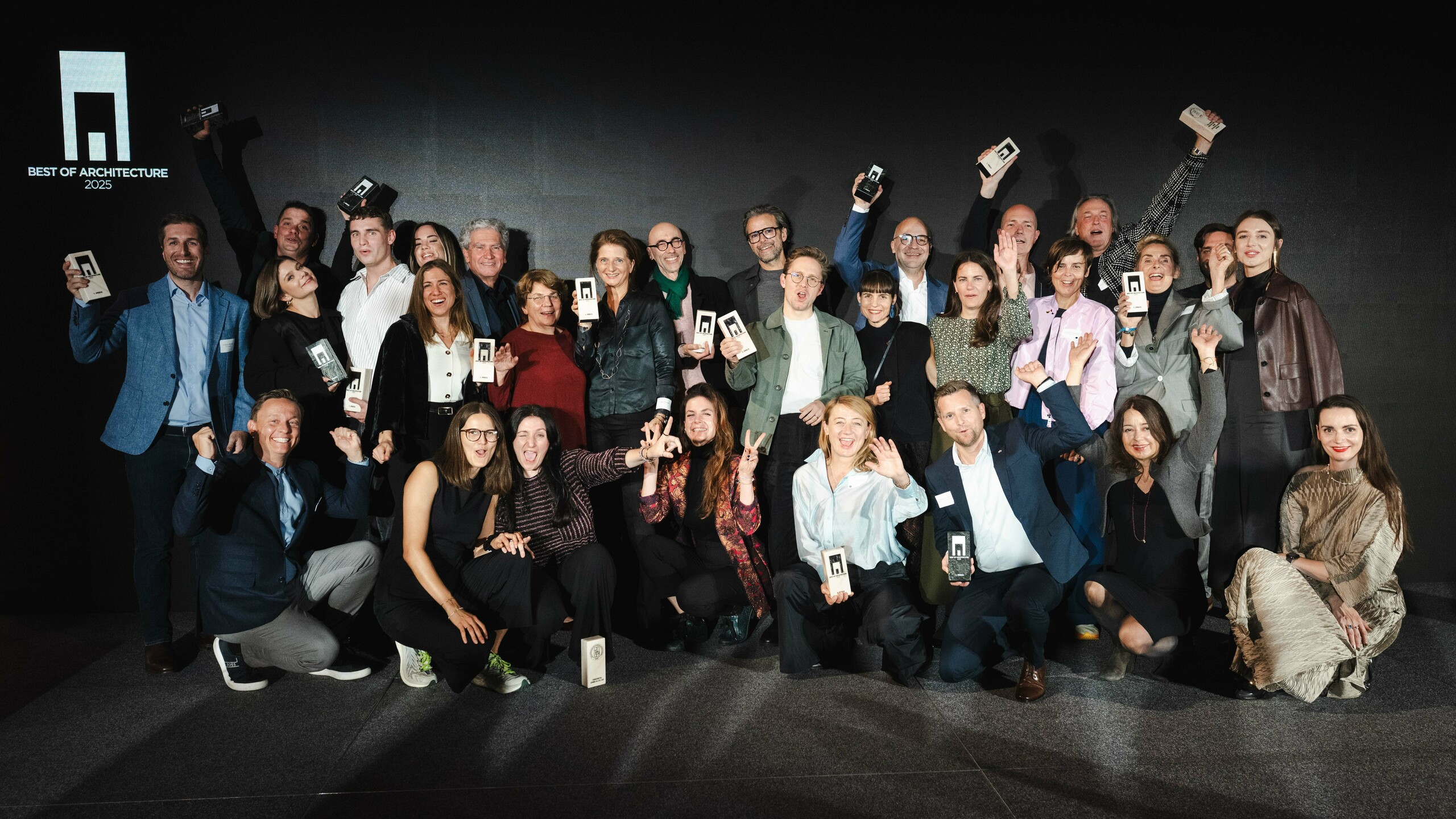 Grupna fotografija pobjednika na dodjeli nagrada „Stambene zgrade godine“ 2025.: dobitnici stoje ispred elegantne crne pozornice s logom „Best of Architecture“ i ponosno drže svoje nagrade. Dinamične poze, moderna odjeća i efektno osvjetljenje naglašavaju važnost događaja, dok scena simbolizira inovaciju i arhitektonsku izvrsnost.