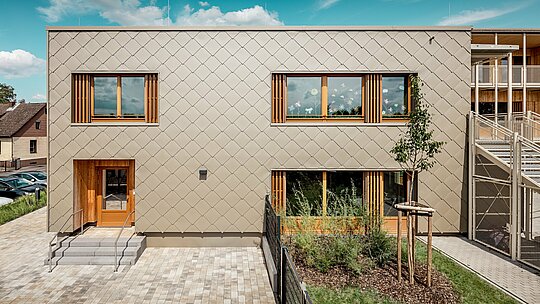 Stražnja strana moderne dječje ustanove u Alzenauu prikazana je iz frontalne perspektive i impresionira jasnim linijama te upečatljivom fasadom od brončano obojenih PREFA zidnih rombova 44 × 44. Geometrijska struktura fasade stvara skladan uzorak „ljuski“, koji je naglašen smirenim, horizontalnim rasporedom prozora. Prostrani drveni okviri i vertikalne lamele unose prirodne naglaske te stvaraju toplu i pozivajuću atmosferu. Promišljena kombinacija dugotrajnog aluminija i lokalnog drva simbolizira održivu arhitekturu visoke estetske kvalitete. Slika ističe preciznu izvedbu, vrhunski odabir materijala i uspješnu integraciju zgrade u urbani kontekst.