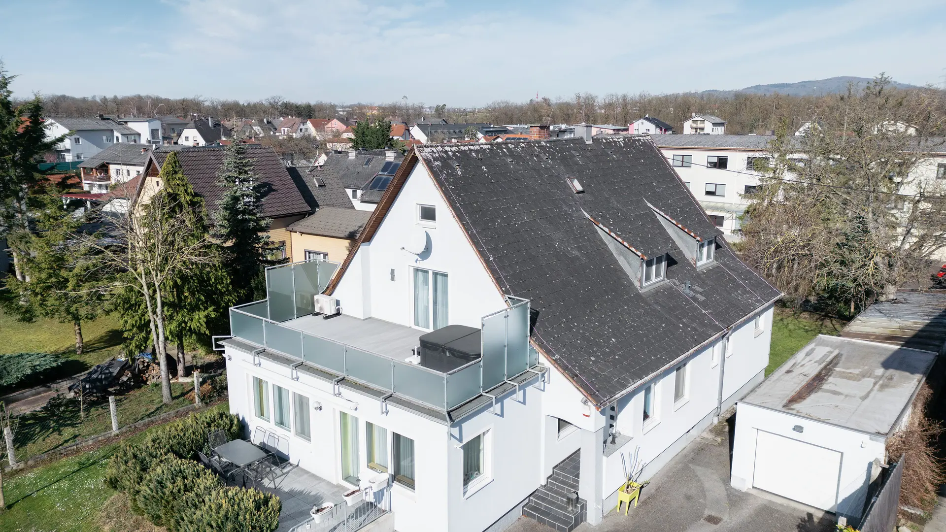 Pogled iz ptičje perspektive na zgradu s starim eternit krovom prekrivenim zemljom i mahovinom. Na krovu nisu vidljivi solarni paneli. Balkon u prvom planu ima staklenu ogradu i nalazi se na posljednjem katu zgrade.