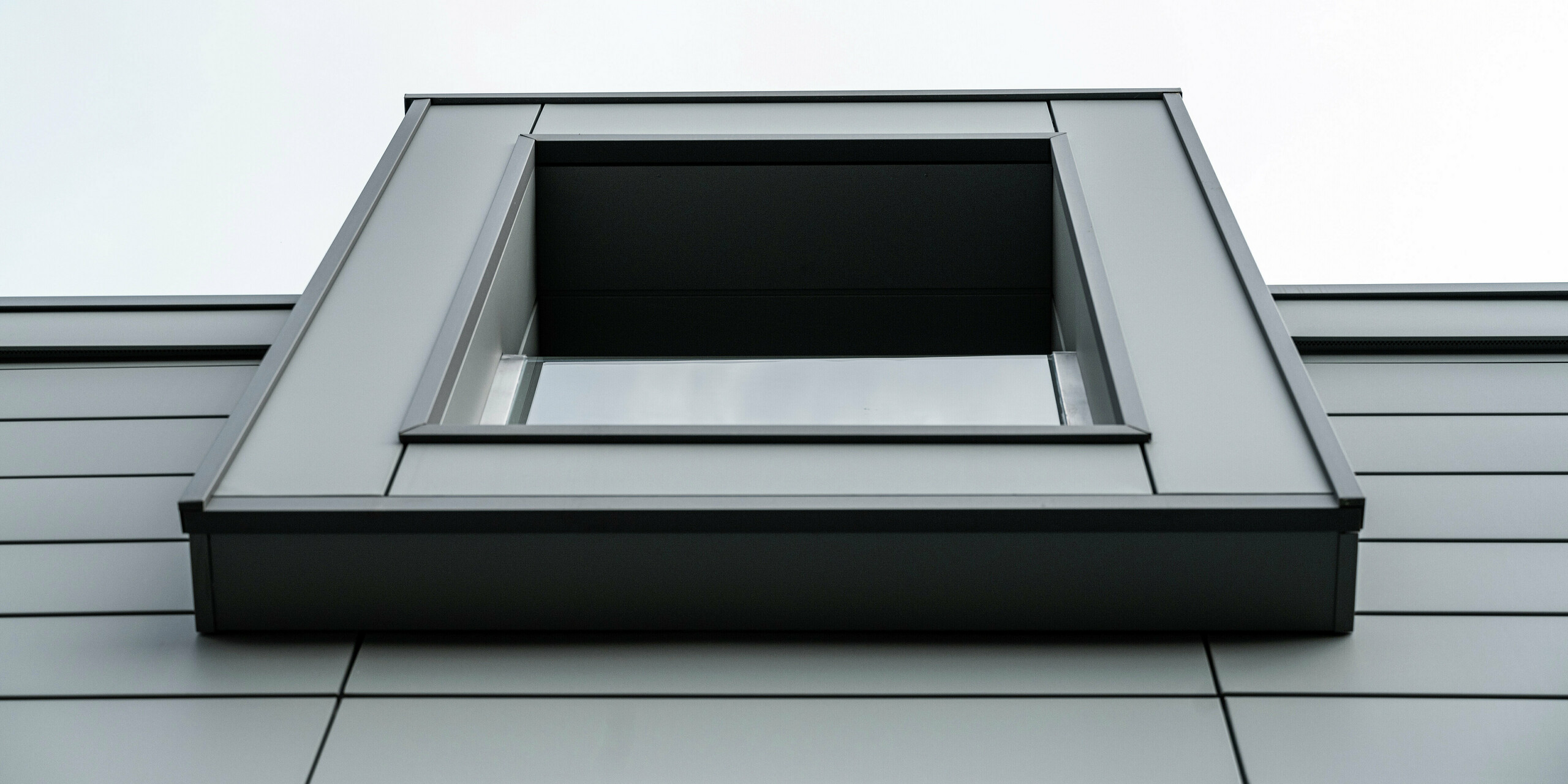 Nahaufnahme von unten auf ein auskragendes Fenster einer Wohnanlage in Faloppio, Italien. Die klare, geometrische Form des Fensters in Kombination mit der langlebigen PREFA Siding Aluminium-Fassade unterstreicht die moderne Architektur und das minimalistische Design des Gebäudes. 
