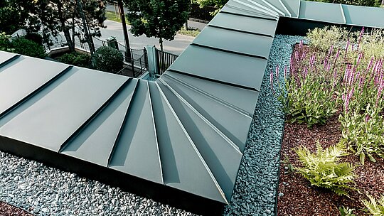 Snimka prikazuje detaljan krupni plan izvedbe atike budimpeštanske višegeneracijske vile, obložene PREFALZ aluminijem u boji P.10 antracit. Lepezasto raspoređeni limeni elementi precizno se spajaju na rubu zgrade i time demonstriraju visoku savitljivost i kvalitetu obrade PREFA aluminijskih proizvoda. Antracitni metal stvara elegantan kontrast prema ozelenjenom krovnom području s šljunčanom podlogom i cvatućim biljkama. Prikazana scena naglašava dugotrajnost, otpornost na vremenske uvjete i tehničku sofisticiranost PREFA rješenja.