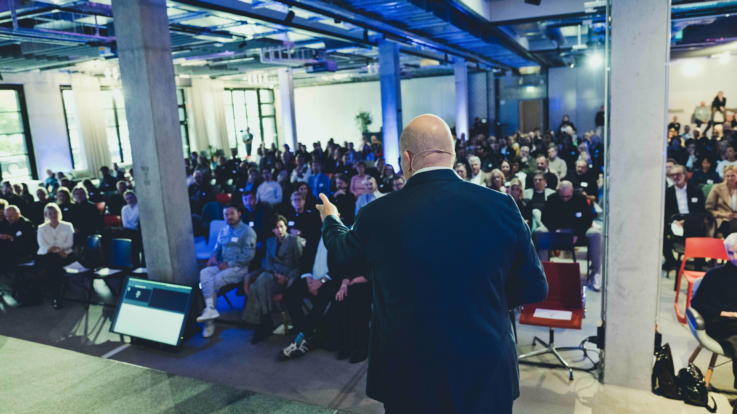 Zanimljiva scena predavanja na dodjeli nagrada „Stambene zgrade godine“ 2025.: pogled s pozornice na potpuno popunjen auditorij u modernom, svjetlom obasjanom prostoru s industrijskim dizajnom. U prvom planu predavač stoji ispred velikog ekrana, dok pažljivi slušaoci prate izlaganje. Događaj naglašava inovacije i aktualne arhitektonske trendove.