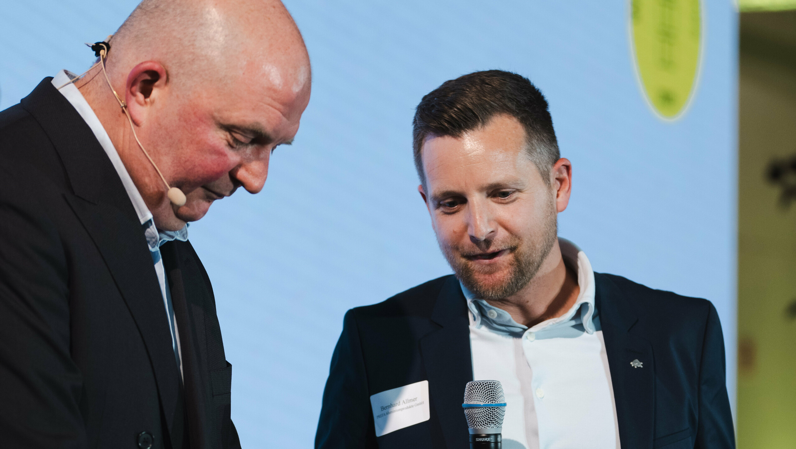 Dodjela nagrade na natječaju „Stambene zgrade godine“ 2025.: dvije osobe su na pozornici, jedna drži mikrofon i nagradu „Architects’ Choice“, dok druga priprema uručenje. Muškarac s mikrofonom i nagradom je Bernhard Allmer, odgovoran za PREFA solarne proizvode. U pozadini svijetli veliki projektorski ekran s logom događaja i zelenim akcentima. Scena simbolizira inovaciju, kvalitetu arhitekture i priznanje PREFALZ solarnom modulu kao održivom rješenju za moderne krovne sustave.