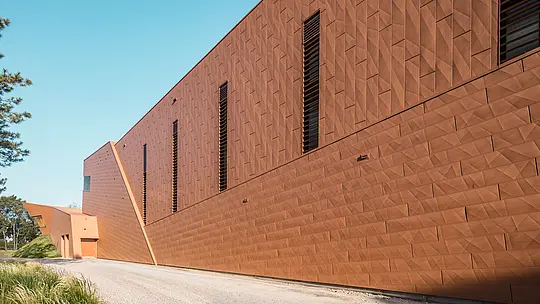 Dinamično presavijena fasada crpne stanice za pitku vodu WML u Lottumu s precizno postavljenim, uskim prozorskim otvorima kao arhitektonskim atraktivnim elementima. Upečatljiva PREFA Siding.X aluminijska fasada u toplom izgledu hrđe stvara živahnu igru svjetla, sjene i geometrije duž jasnog ruba volumena zgrade. Izvanredna struktura povezuje tehničku funkciju sa skulpturalnom estetikom. Lagani, dugotrajni i potpuno reciklabilni PREFA aluminijski paneli odlikuju se visokom otpornošću na vremenske uvjete, stabilnošću oblika te održivim fasadnim omotačem koji zahtijeva minimalno održavanje.