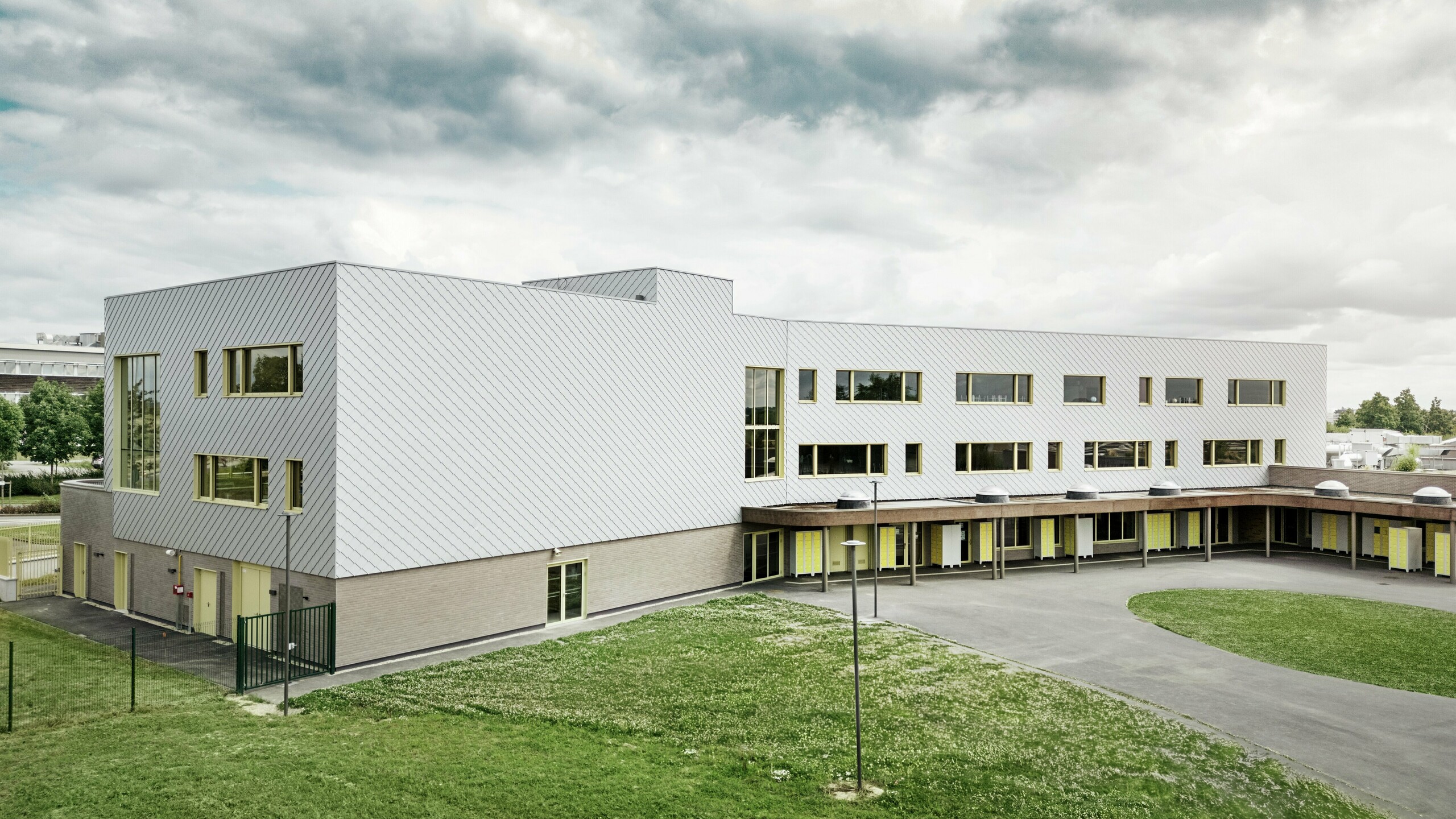 Slika prikazuje stražnju stranu Collège Léontine Dolivet. U prvom planu nalazi se prostrano asfaltirano školsko dvorište s jasnim linijama i otvorenim površinama, pogodnim za odmore i slobodne aktivnosti. Nekoliko zelenih površina razbija monotoniju okoline. U pozadini se uzdiže moderna školska zgrada s upečatljivom aluminijskom fasadom od srebrno-metalnih PREFA zidnih rombova, koja reflektira svjetlo i stvara dinamičan efekt. Više velikih prozora osigurava svjetlost u unutrašnjosti. Scena je snimljena danju, s lagano oblačnim nebom, što posebno naglašava metalnu površinu.