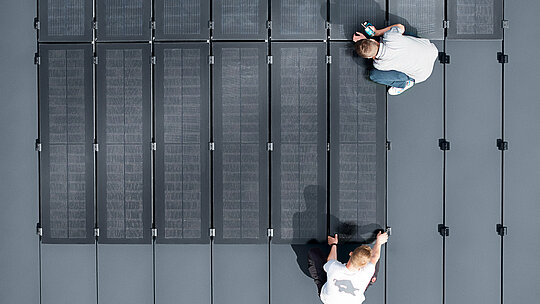  Dvoje ljudi instalira PREFALZ solarne module na velikoj sivoj površini krova, koja je prekrivena panelima s dvostrukim stojećim šavom.