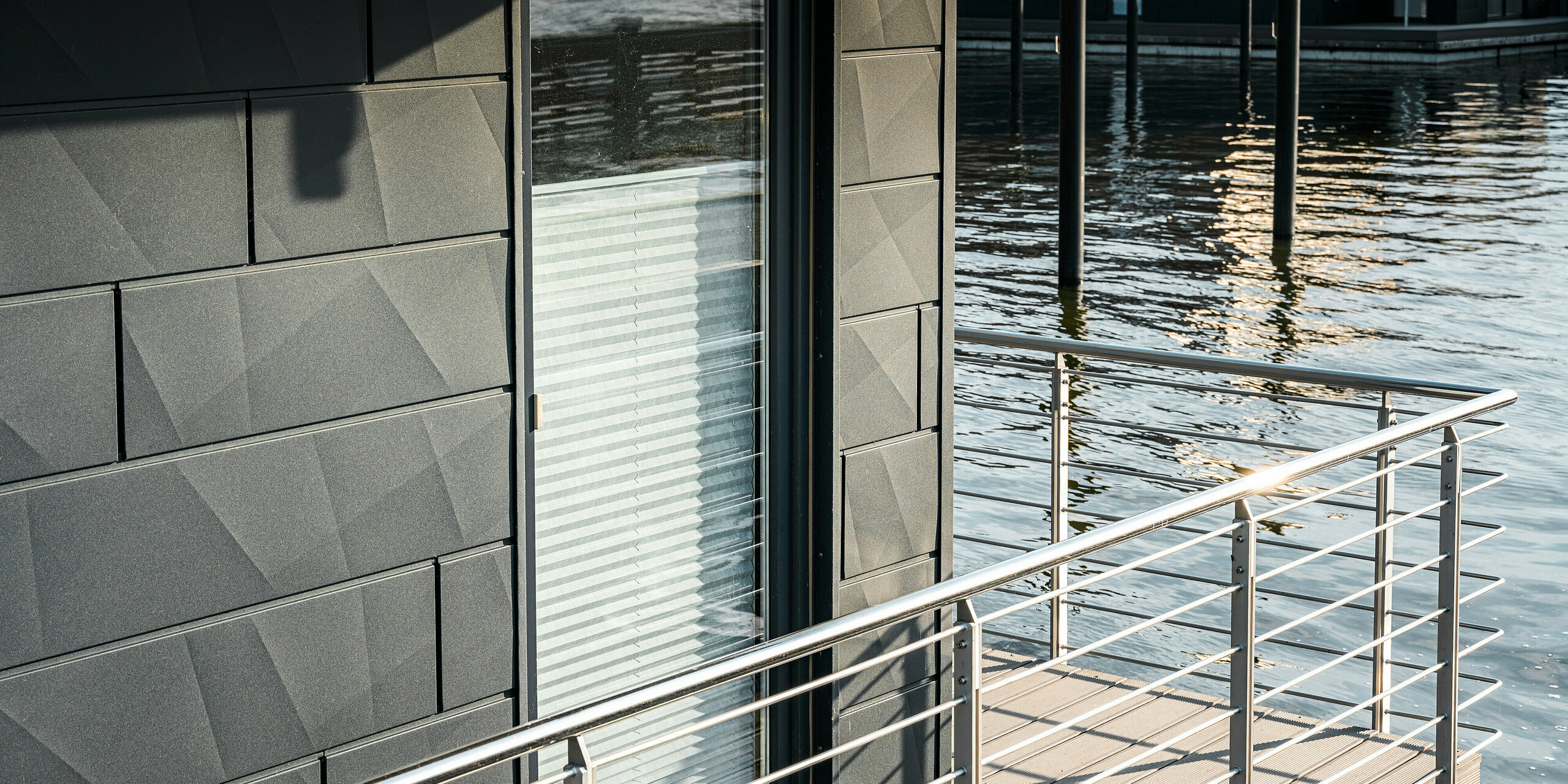 Detailansicht der texturierten Oberfläche einer PREFA Siding.X Aluminiumfassade in P.10 Anthrazit an einem Wasserhaus im Pionierhafen Schleswig. Die Fassade fängt das Licht der untergehenden Sonne ein, was das Wasser darunter reflektiert und für eine beruhigende Atmosphäre sorgt. Die makellose Verarbeitung und das moderne Geländer zeigen die hohe Qualität und das ästhetische Bewusstsein, das in die Gestaltung dieser nachhaltigen Wohnräume am Wasser eingeflossen ist.