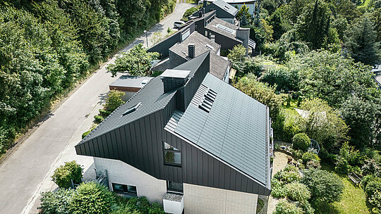 Kosi prikaz moderne obiteljske kuće u Meschedeu, Njemačka. Pomaknuti krov prekriven je PREFALZ-om u P.10 crnoj boji. Aluminijski krov služi kao vizualni naglasak i pruža dugotrajnu zaštitu zgradi. Precizno dvostruko falcno pokrivanje proteže se od krova preko fasade do pročelja. Okružena bujnim zelenilom i mirnom ulicom, energetski učinkovita kuća s inovativnim PREFALZ solarnim modulima savršeno se uklapa u prirodno okruženje.