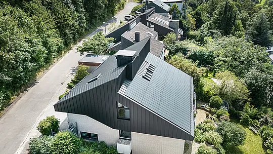 Kosi prikaz moderne obiteljske kuće u Meschedeu, Njemačka. Pomaknuti krov prekriven je PREFALZ-om u P.10 crnoj boji. Aluminijski krov služi kao vizualni naglasak i pruža dugotrajnu zaštitu zgradi. Precizno dvostruko falcno pokrivanje proteže se od krova preko fasade do pročelja. Okružena bujnim zelenilom i mirnom ulicom, energetski učinkovita kuća s inovativnim PREFALZ solarnim modulima savršeno se uklapa u prirodno okruženje.