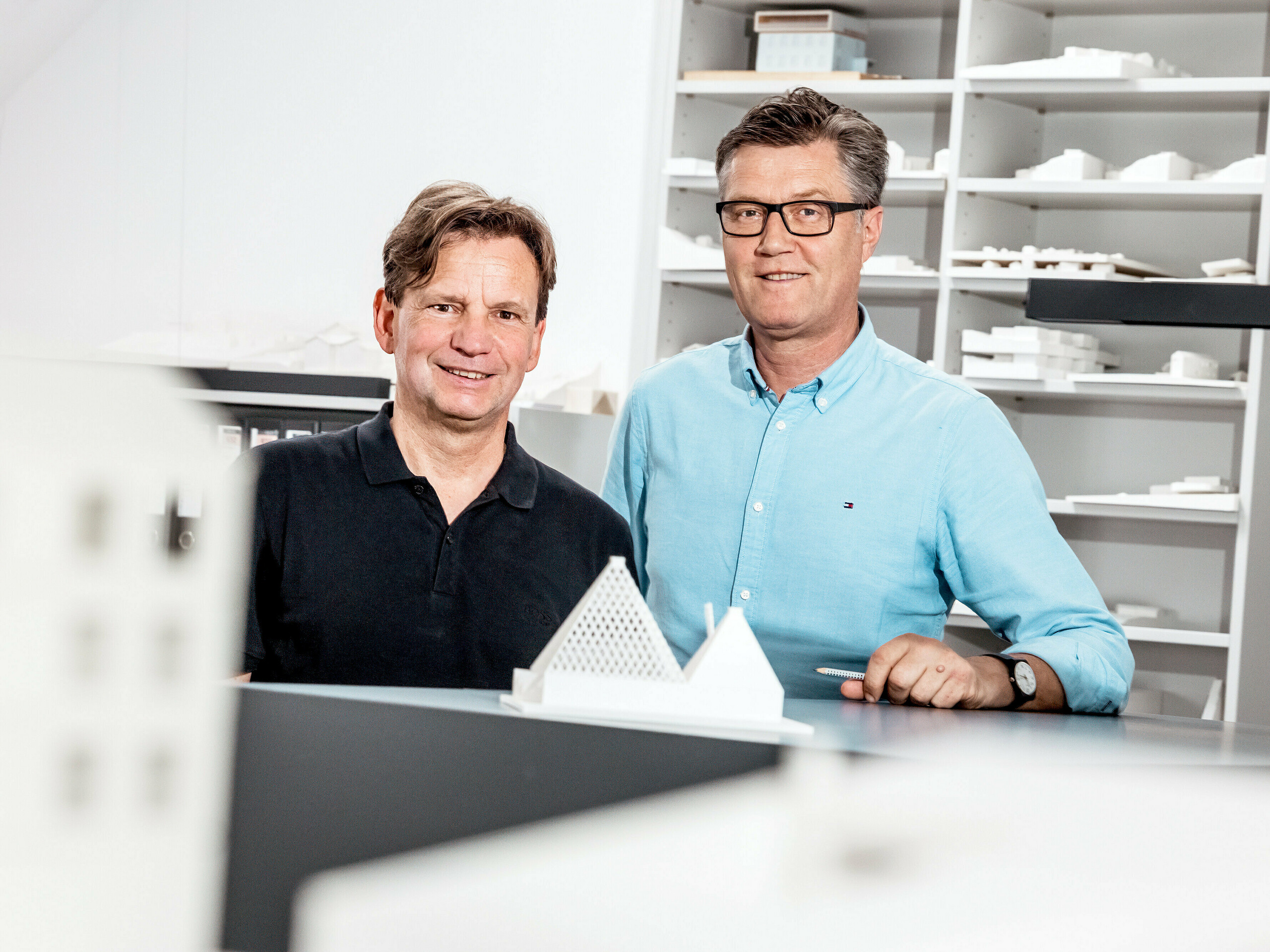 Na slici su arhitekti iz Schneider Lengauer Pühringer, kako ponosno poziraju u svom uredu ispred zbirke arhitektonskih modela. Osmislili su modernu obiteljsku kuću u Freistadtu pod nazivom projekta One Shade of White i poznati su po svom inovativnom i estetskom pristupu arhitekturi. Modeli u pozadini odražavaju njihove kreativne koncepte i pokazuju njihovu sposobnost da moderne dizajnerske ideje spoje s funkcionalnim rješenjima. Obojica nose ležernu odjeću, što naglašava opuštenu, ali istovremeno profesionalnu atmosferu u njihovom radnom okruženju.