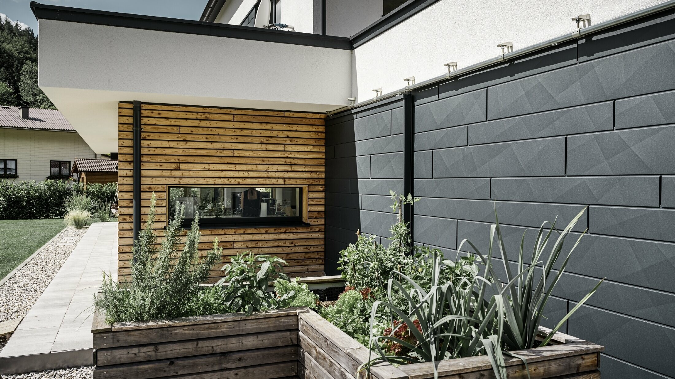 Ova slika prikazuje krupni plan samostojeće kuće, s naglaskom na oblogu pročelja izrađenu od PREFA Siding.X u boji P.10 Anthracite. Tamni paneli pročelja, sa svojom prepoznatljivom naboranom završnom obradom, pružaju privlačan vizualni kontrast živopisnim, drvenim obloženim površinama i okolnom zelenilu. Povišene gredice, posađene raznim biljkama i grmljem, postavljene su ispred aluminijske fasade, naglašavajući prirodni i održivi karakter kuće. Kombinacija modernih materijala i integracija u vrtni prostor prenosi sliku suvremene arhitekture koja se skladno uklapa u svoje okruženje.