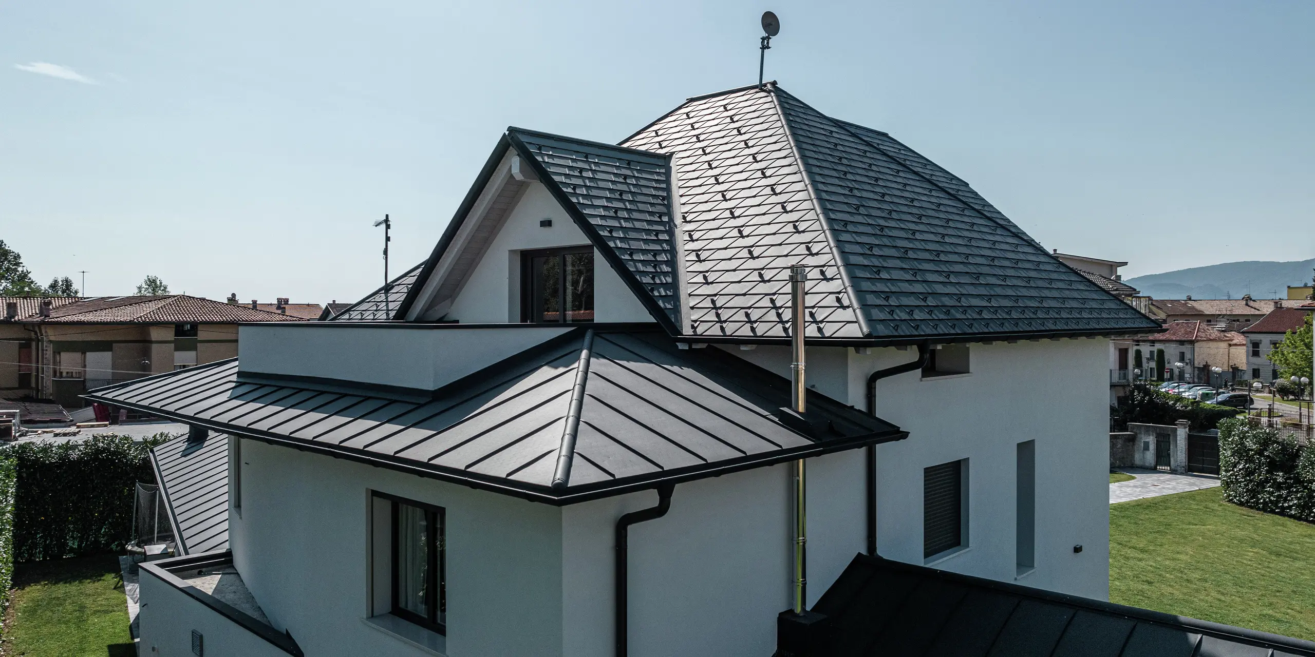 Detailansicht der Dachrückseite eines modernen Einfamilienhauses in Marano Vicentino, Italien. Das Hauptdach ist mit PREFA Dachschindeln in P.10 Anthrazit eingedeckt, während die beiden Anbauten mit einer eleganten Doppelstehfalzdeckung aus Aluminium versehen sind. Die Kombination hochwertiger Aluminiumprodukte sorgt für eine langlebige, witterungsbeständige und ästhetische Dachlösung.