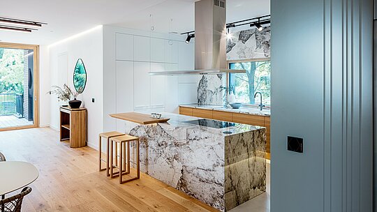 Snimka prikazuje elegantan interijer budimpeštanske višegeneracijske vile s modernom, otvorenom kuhinjom. Upečatljivi elementi su mramorirane površine prirodnog kamena na kuhinjskom otoku i stražnjoj stijeni, u kombinaciji s toplim akcentima hrastovine i visokokvalitetnim ugradbenim elementima. Prostrane staklene površine propuštaju obilje dnevnog svjetla i skladno povezuju prostor s okolnim zelenilom. Jasna linijska struktura, plemenit odabir materijala i precizna izvedba odražavaju visoke arhitektonske standarde objekta, čiji je vanjski izgled nadopunjen dugotrajnim PREFA aluminijskim proizvodima.